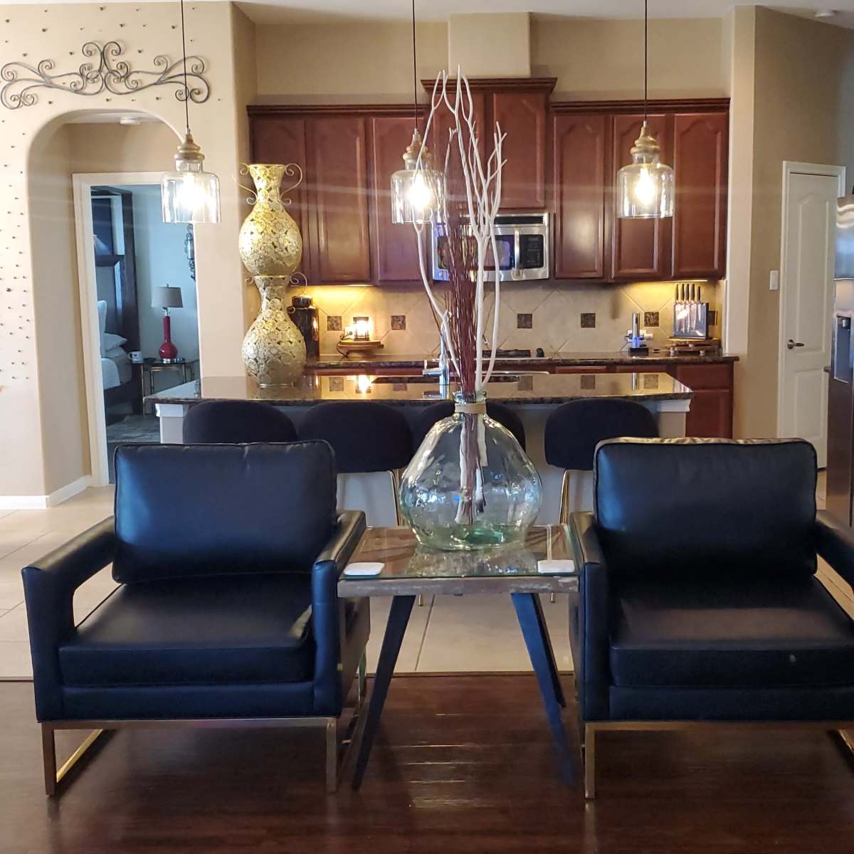 A modern living space with two black armchairs facing a small table, which features a decorative vase, and a kitchen area with brown cabinets and stainless steel appliances in the background.