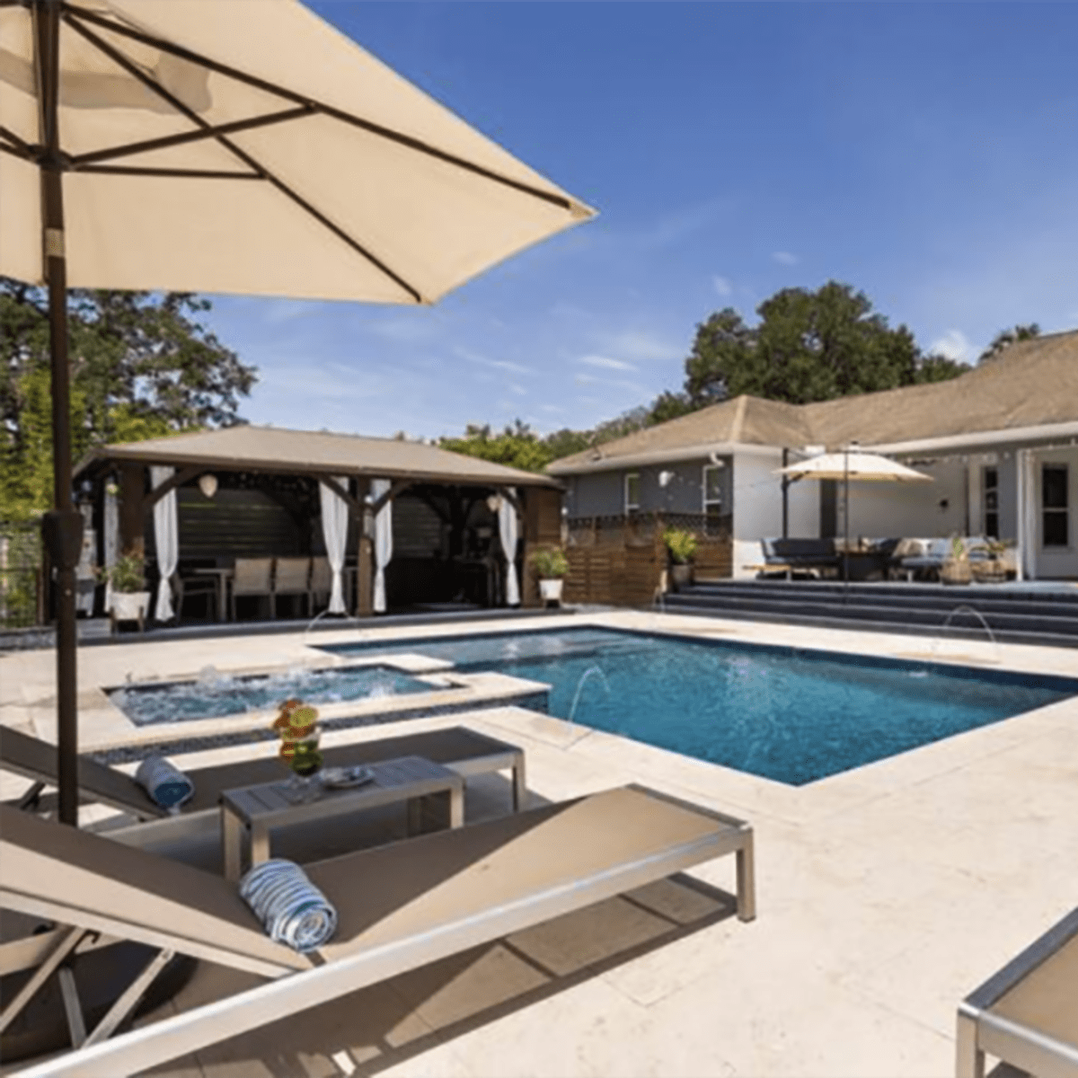 A backyard area featuring a rectangular swimming pool surrounded by lounge chairs, sun umbrellas, and outdoor seating under a gazebo.