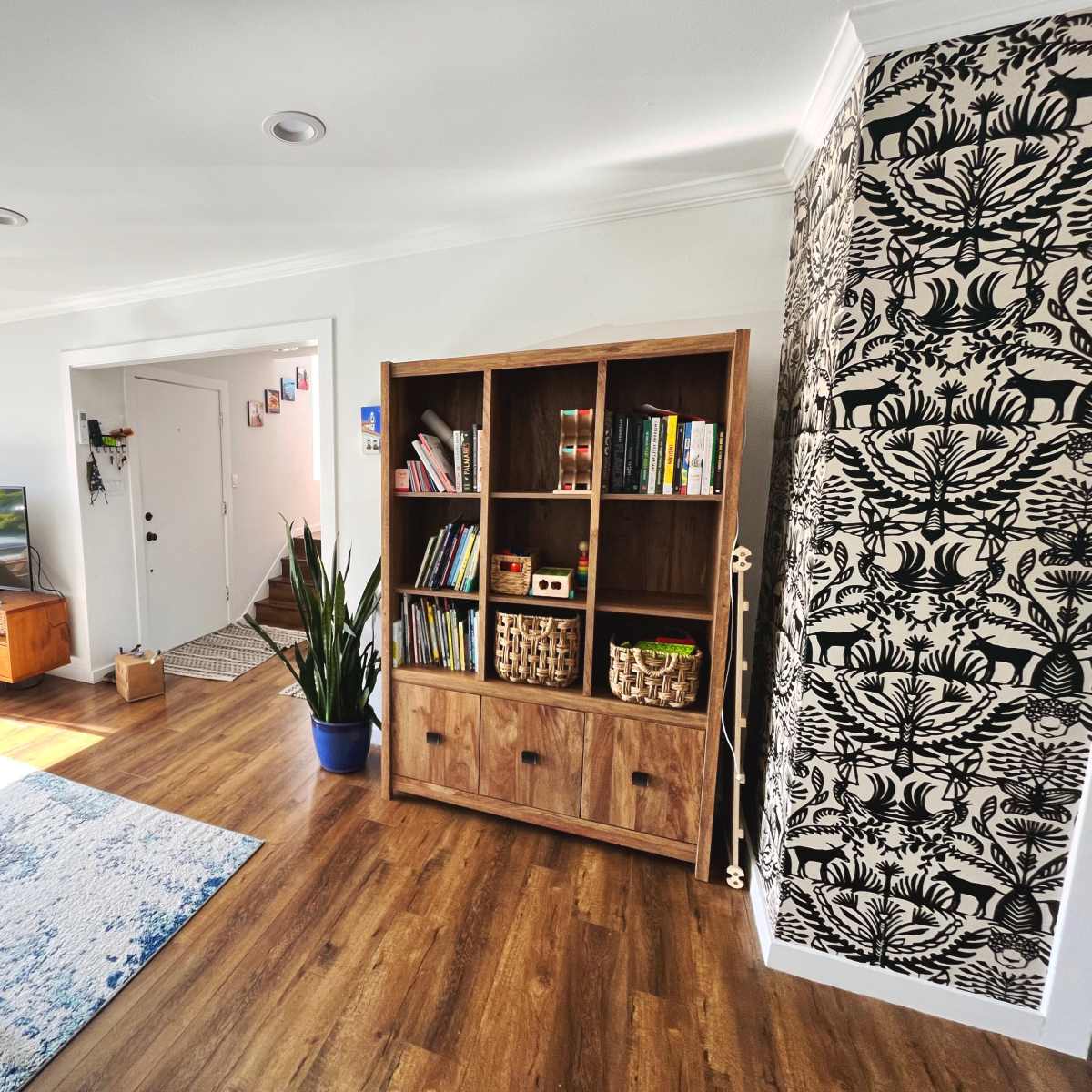 The living room features a large bookshelf, a television stand, and a door leading outside, with a patterned wallpaper accent wall.