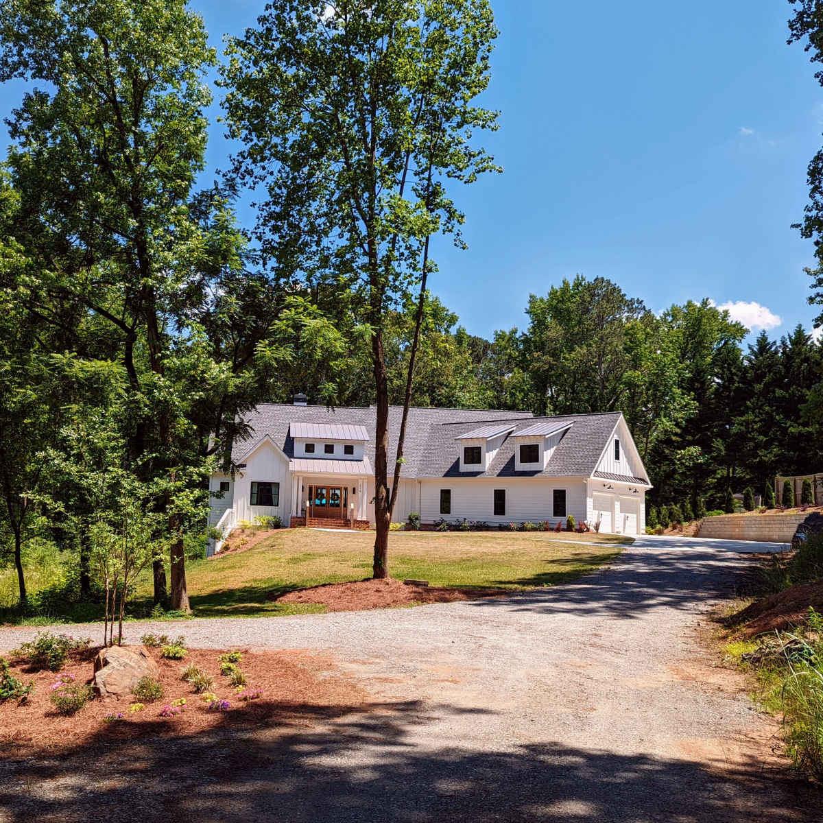 A modern white house with a sloped roof is set among trees, accessed by a gravel driveway.