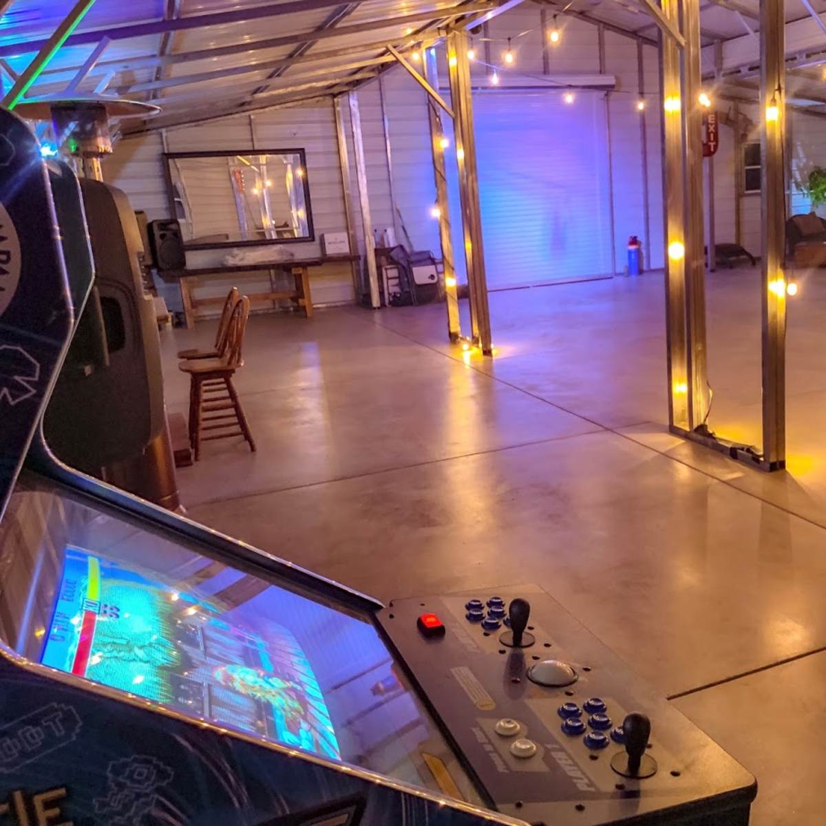 A spacious indoor area with a video game arcade machine in the foreground, string lights illuminating the space, and a white sofa against a wooden backdrop.