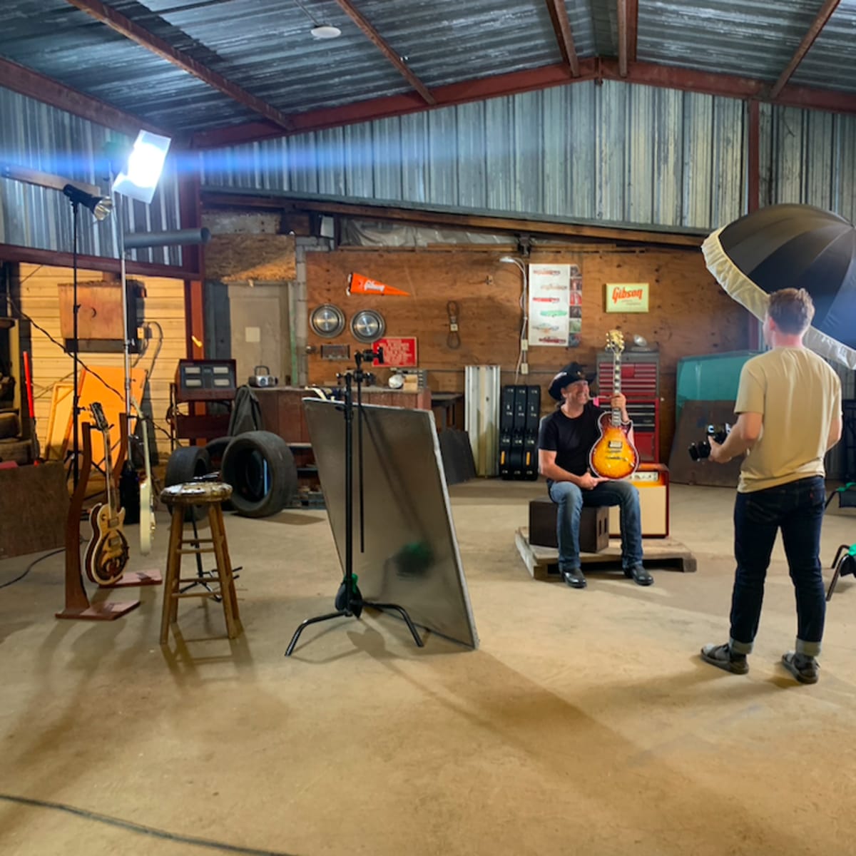 A person sits on a stool with an electric guitar in a dimly lit workshop, while another person adjusts lighting equipment nearby.