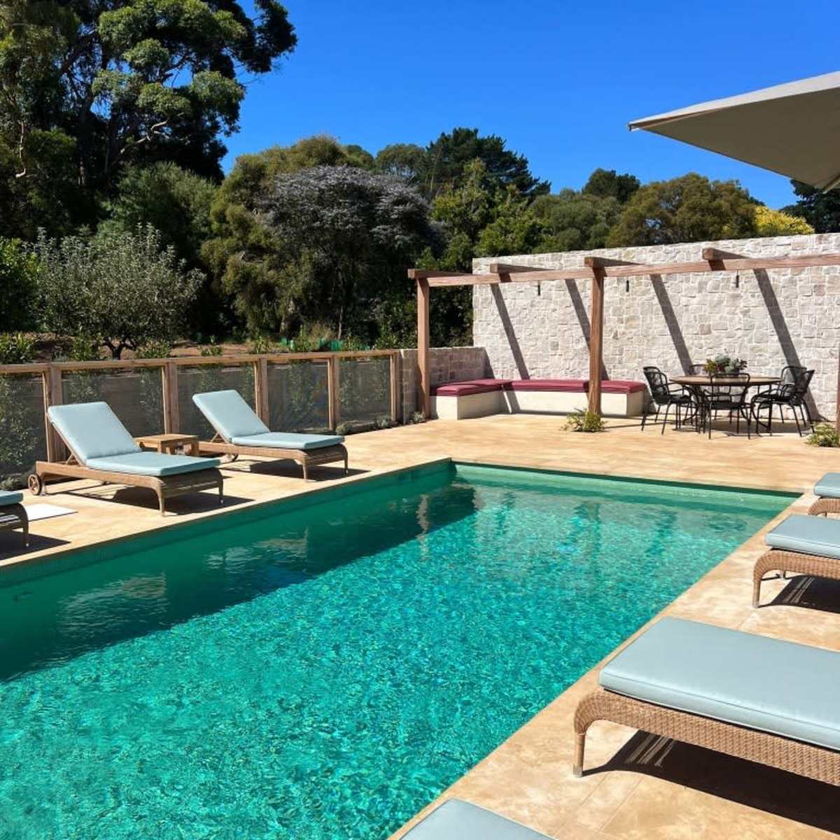 A swimming pool surrounded by lounge chairs, with a dining area and landscaping in the background.