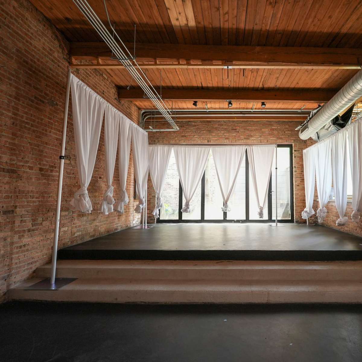 A spacious room with exposed brick walls, wooden ceilings, and white drapes hanging from a pole near large windows.