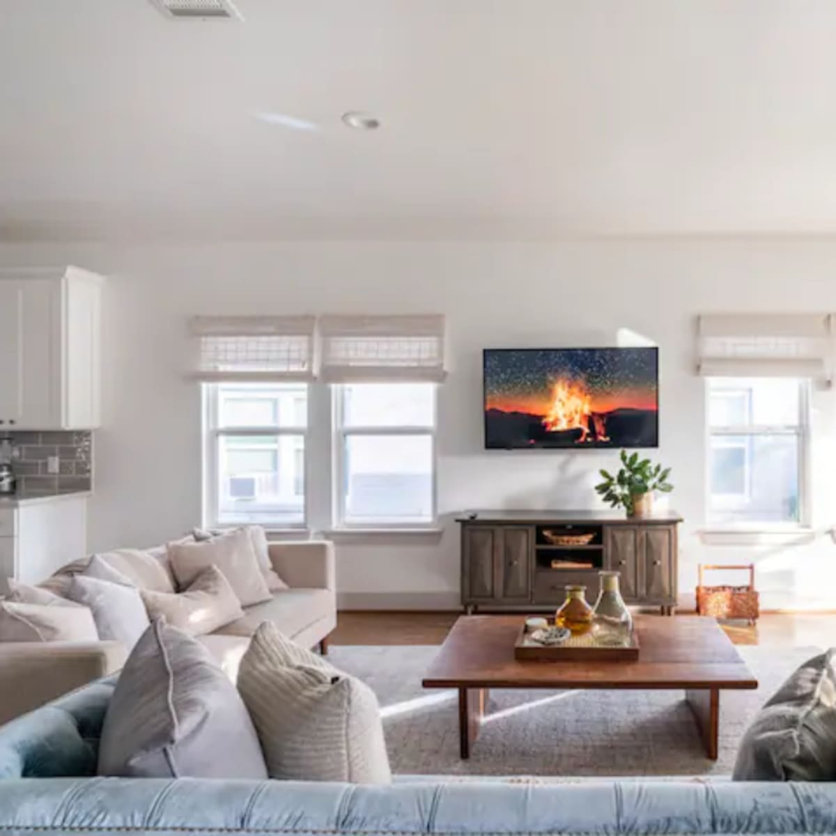 A living room features a cozy arrangement of sofas and a central coffee table, with a television displaying a campfire scene on the wall above a wooden console.