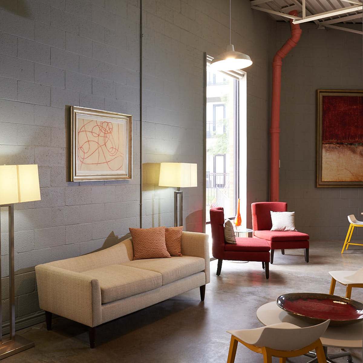 A modern lounge area with a beige sofa, red accent chairs, and floor lamps against a gray wall, illuminated by natural light through large windows.