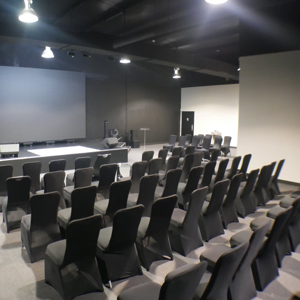 A spacious auditorium features a large projection screen at the front and rows of black chairs facing it.