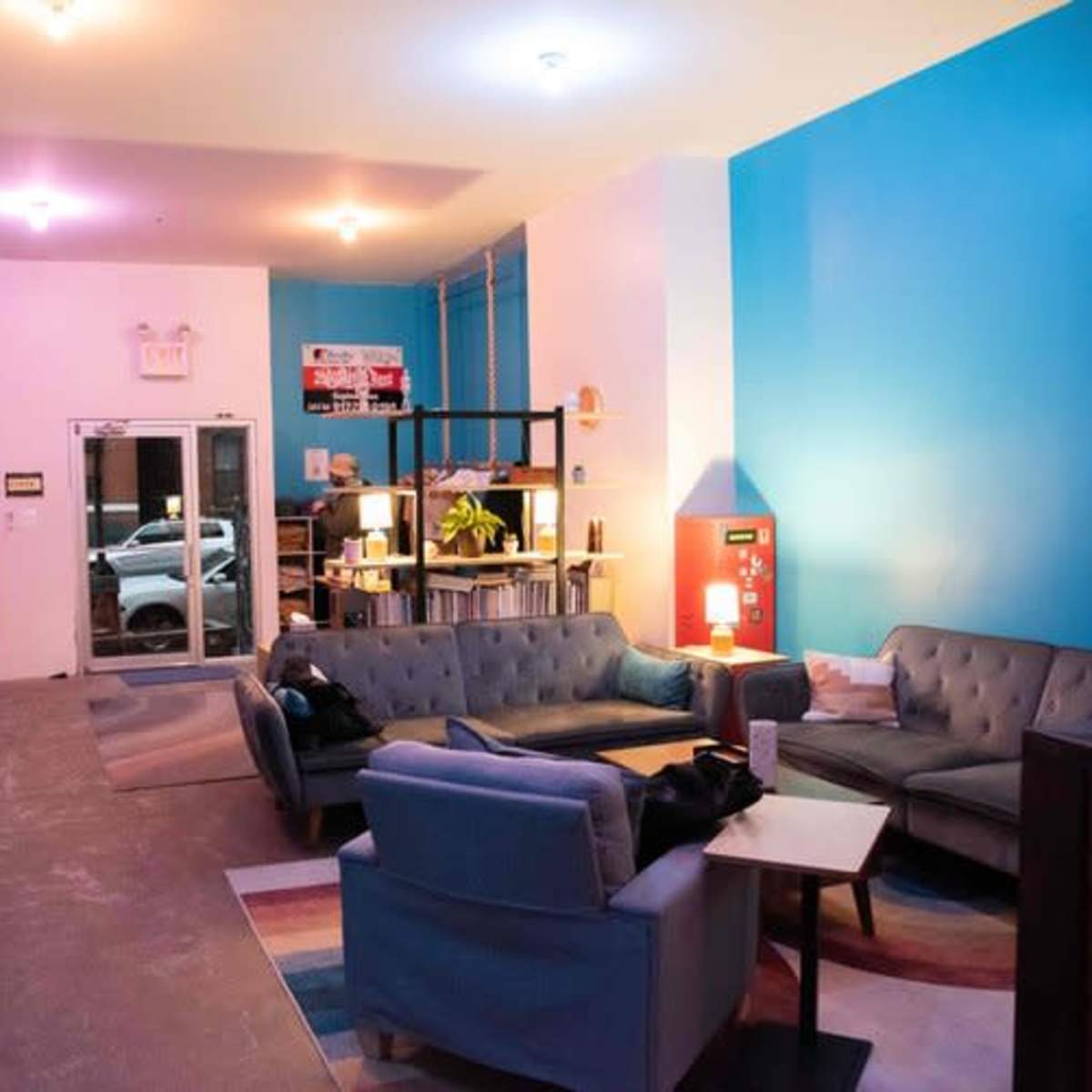 A spacious lounge area with blue walls, various seating arrangements, and a large window facing the street.