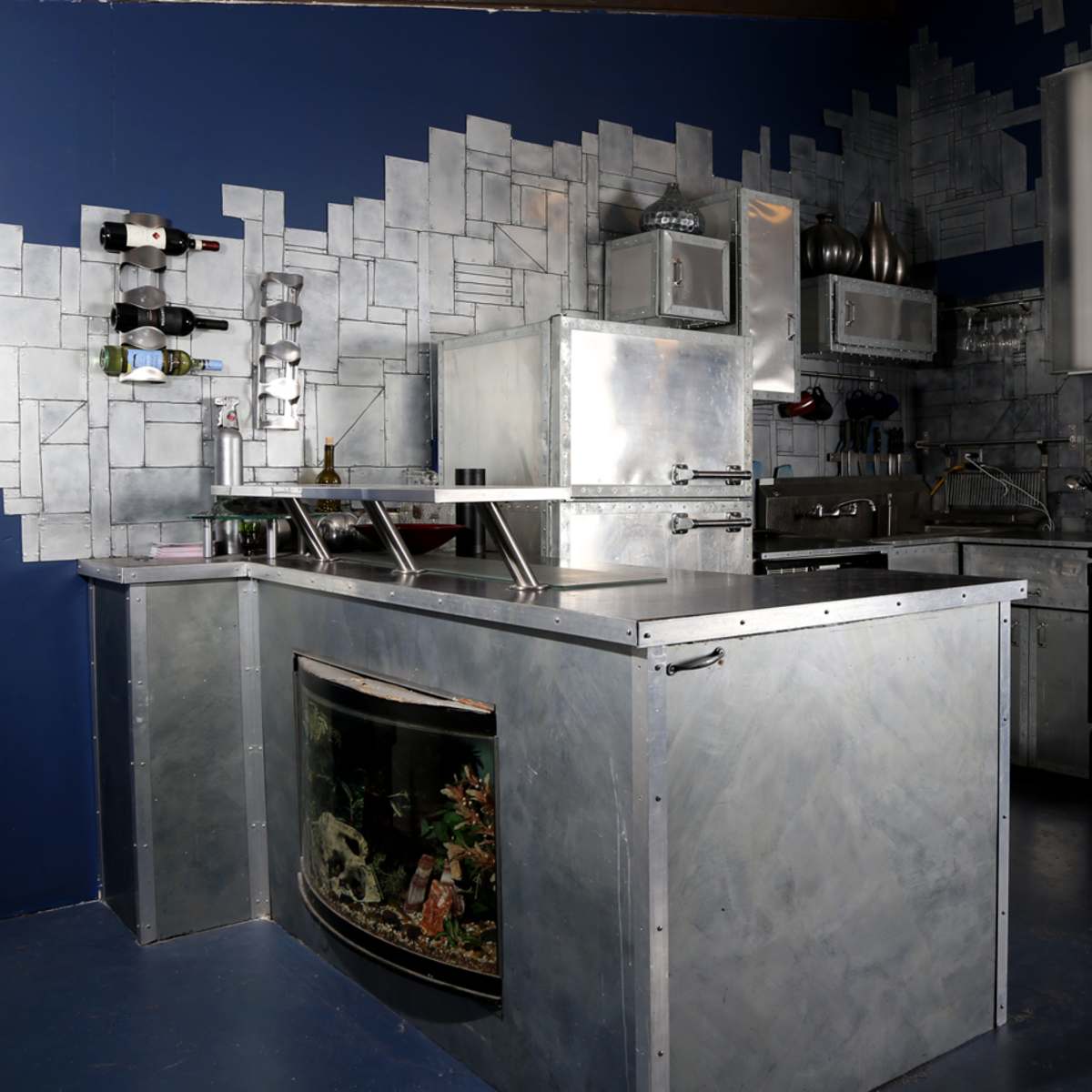 The kitchen features metallic surfaces, an aquarium integrated into the counter, and modern appliances against a geometric wall design.