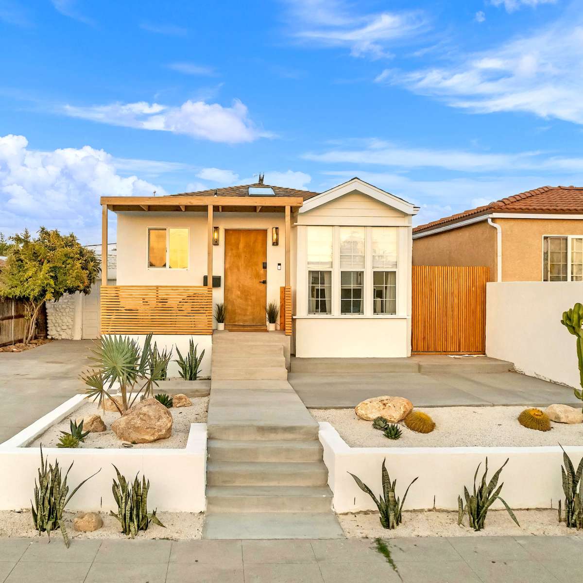 A single-story house with a wooden porch, surrounded by gravel landscaping and desert plants, is set against a blue sky with clouds.
