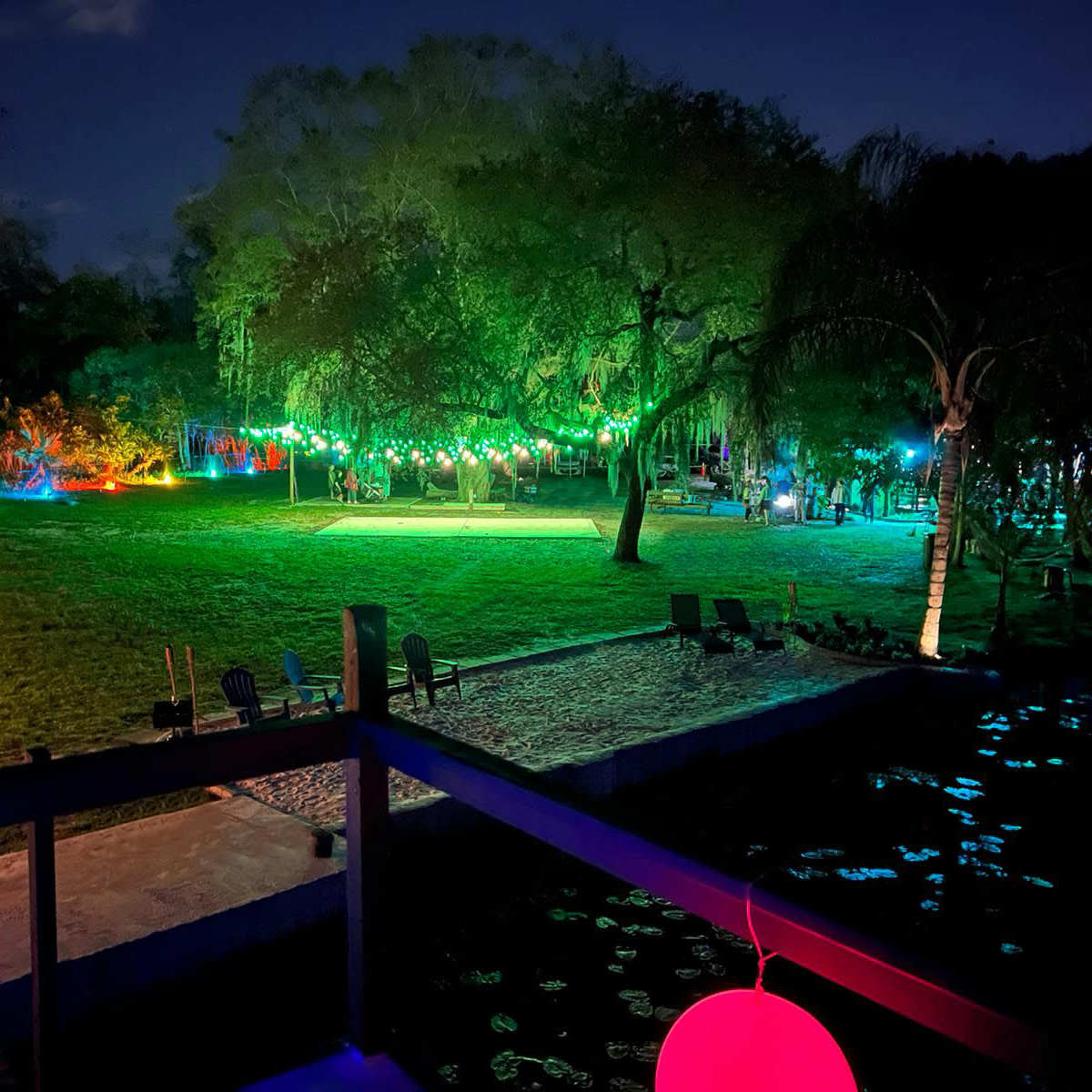 A night scene of a landscaped area illuminated by colorful lights with a grassy field and a waterfront.