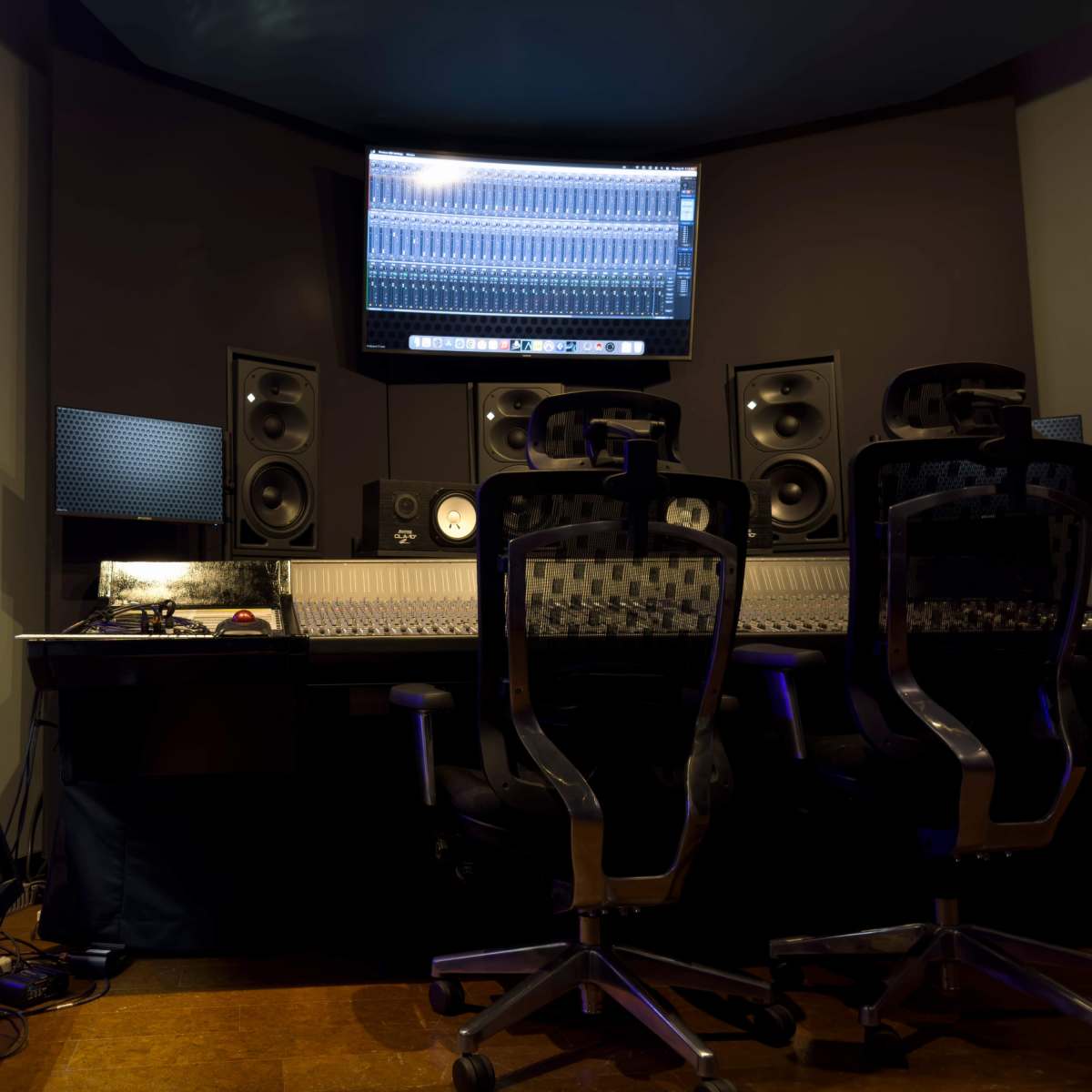 A music recording studio with audio mixing equipment, speakers, and two ergonomic chairs positioned in front of a large mixing console and a wall-mounted monitor.
