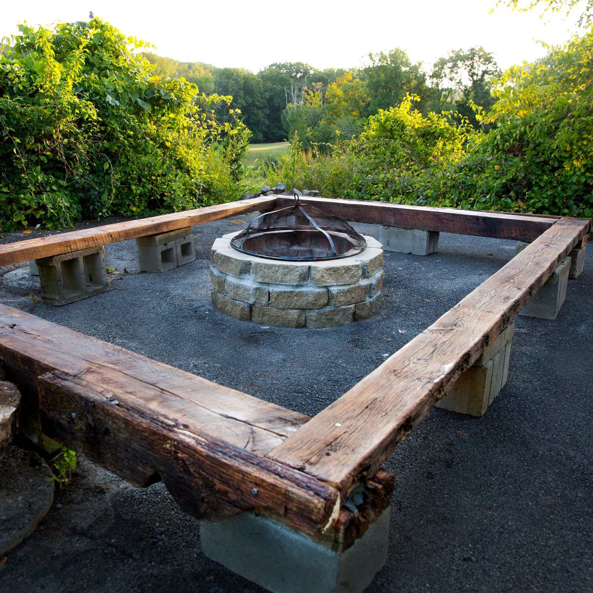 A rectangular fire pit surrounded by wooden seating and stone blocks, set in a natural landscape.