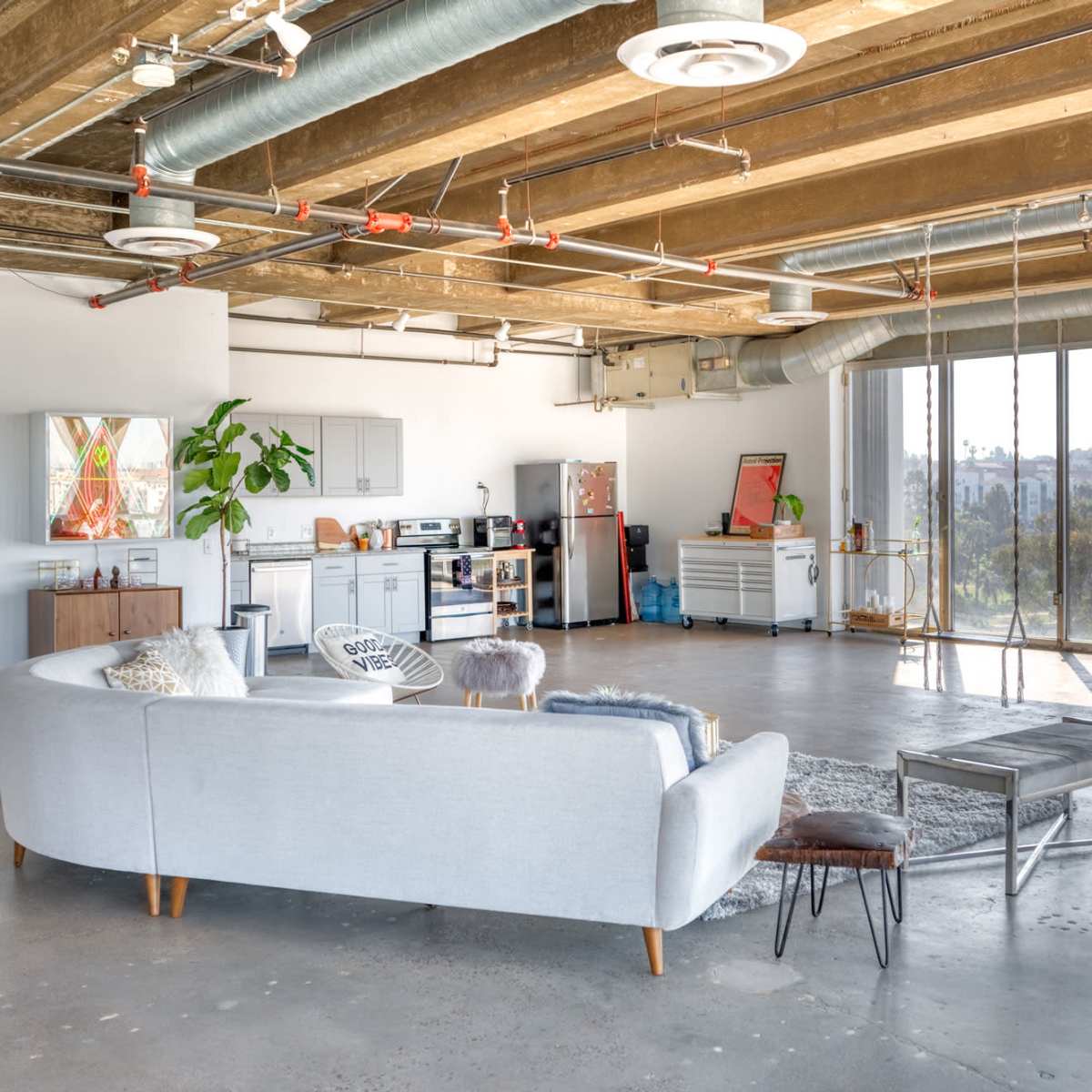 A spacious, modern living room with industrial-style ceilings, a gray sectional sofa, and large windows overlooking a view of the skyline.
