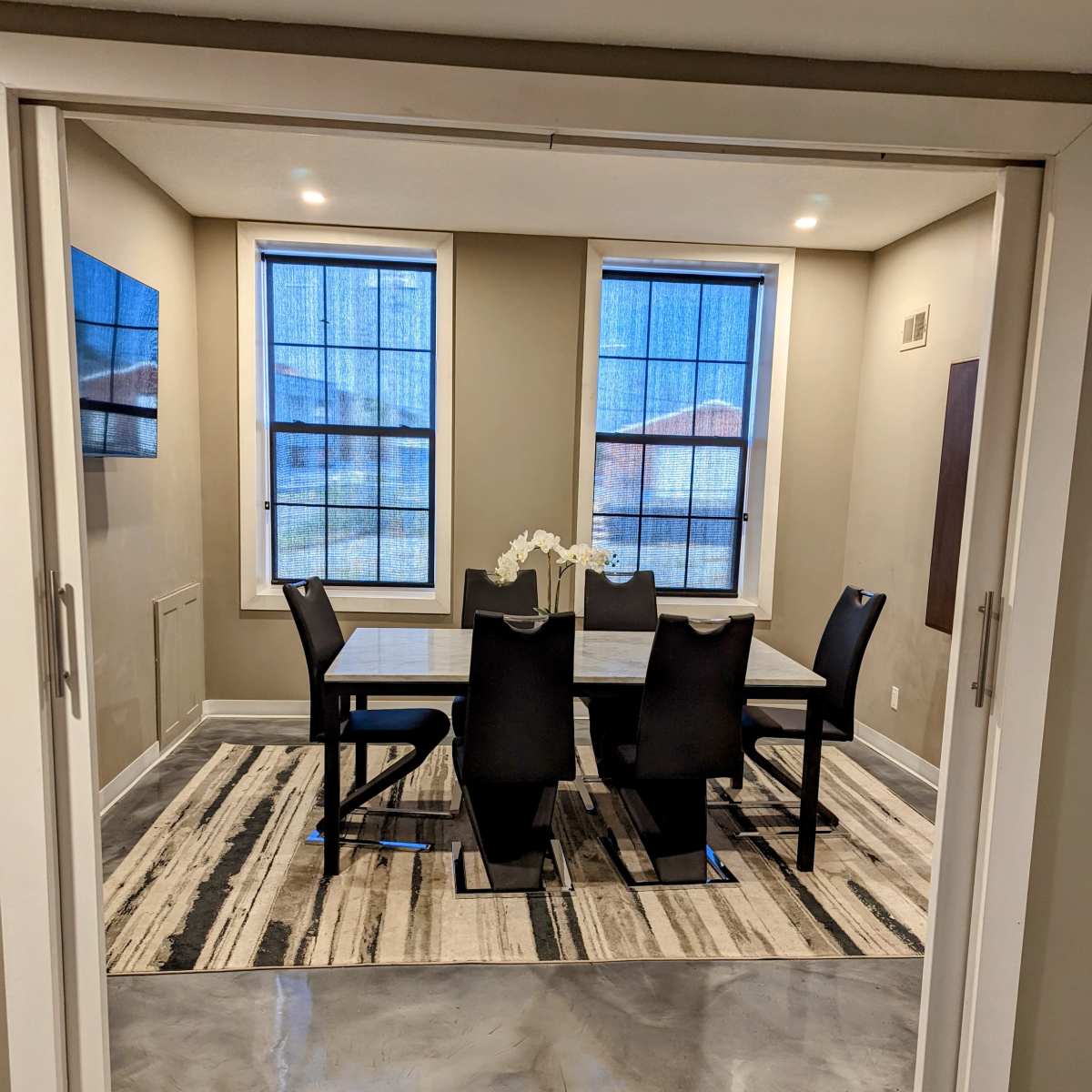 A dining room features a rectangular table surrounded by six black chairs, with large windows illuminating the space and a concrete floor.