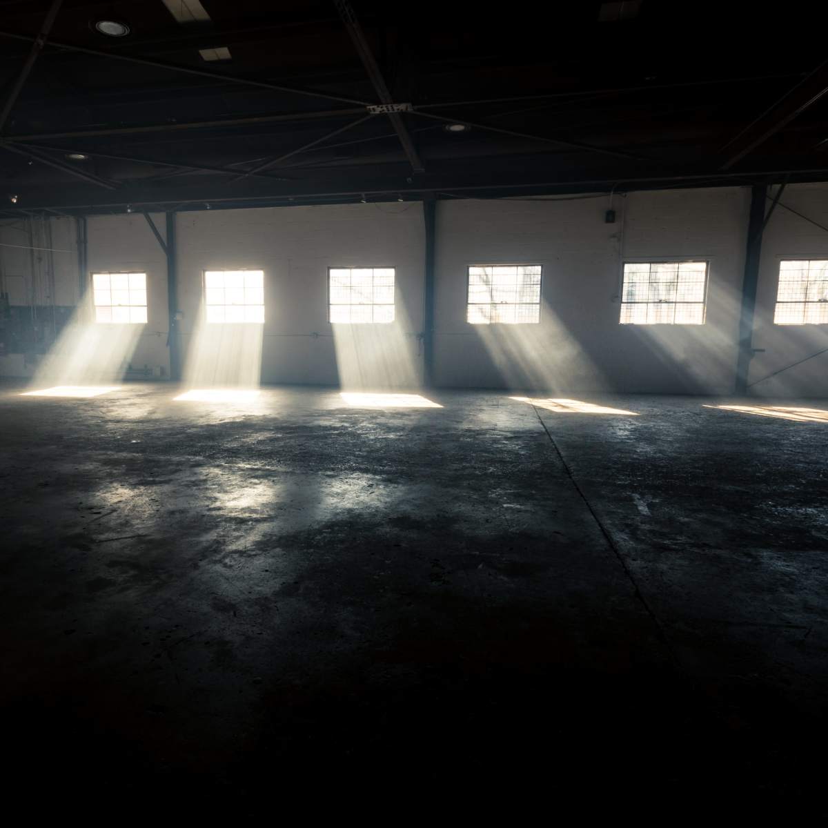 Light streams through windows, casting beams onto the empty, concrete floor of a warehouse.