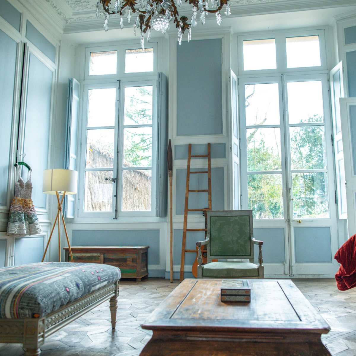 A bright living room features large windows, vintage furniture, and a wooden coffee table, with a cozy atmosphere created by natural light.