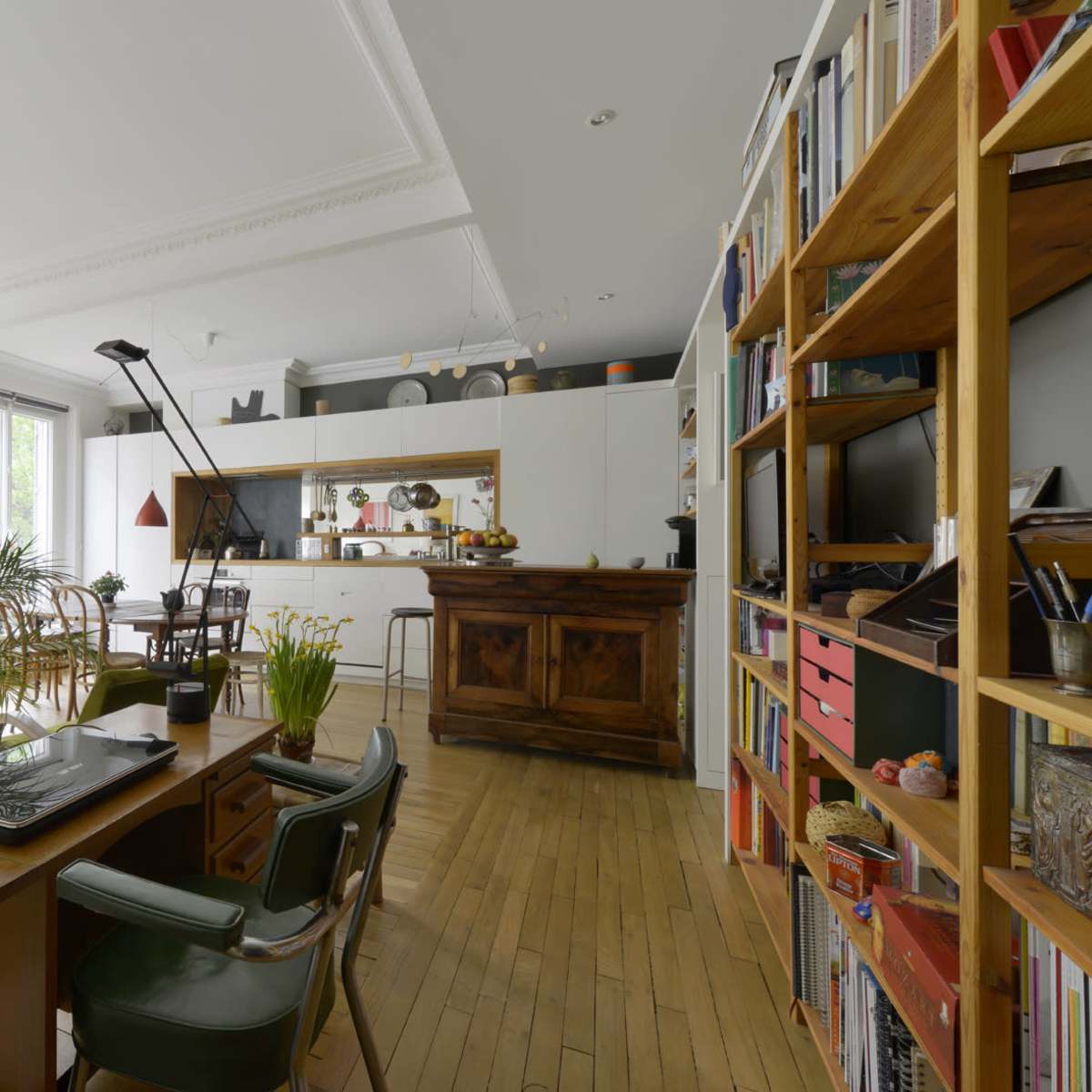 A modern living space features a wooden dining table, a kitchen area with white cabinetry, and a wooden bookshelf filled with books, plants, and decorative items.