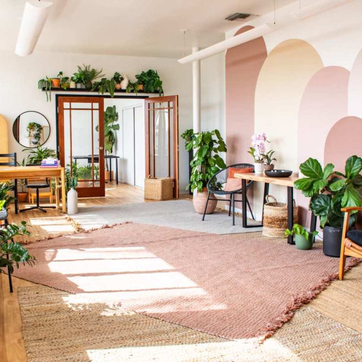 A bright, modern interior space with wooden floors, various plants, a dining area, and colorful wall designs.