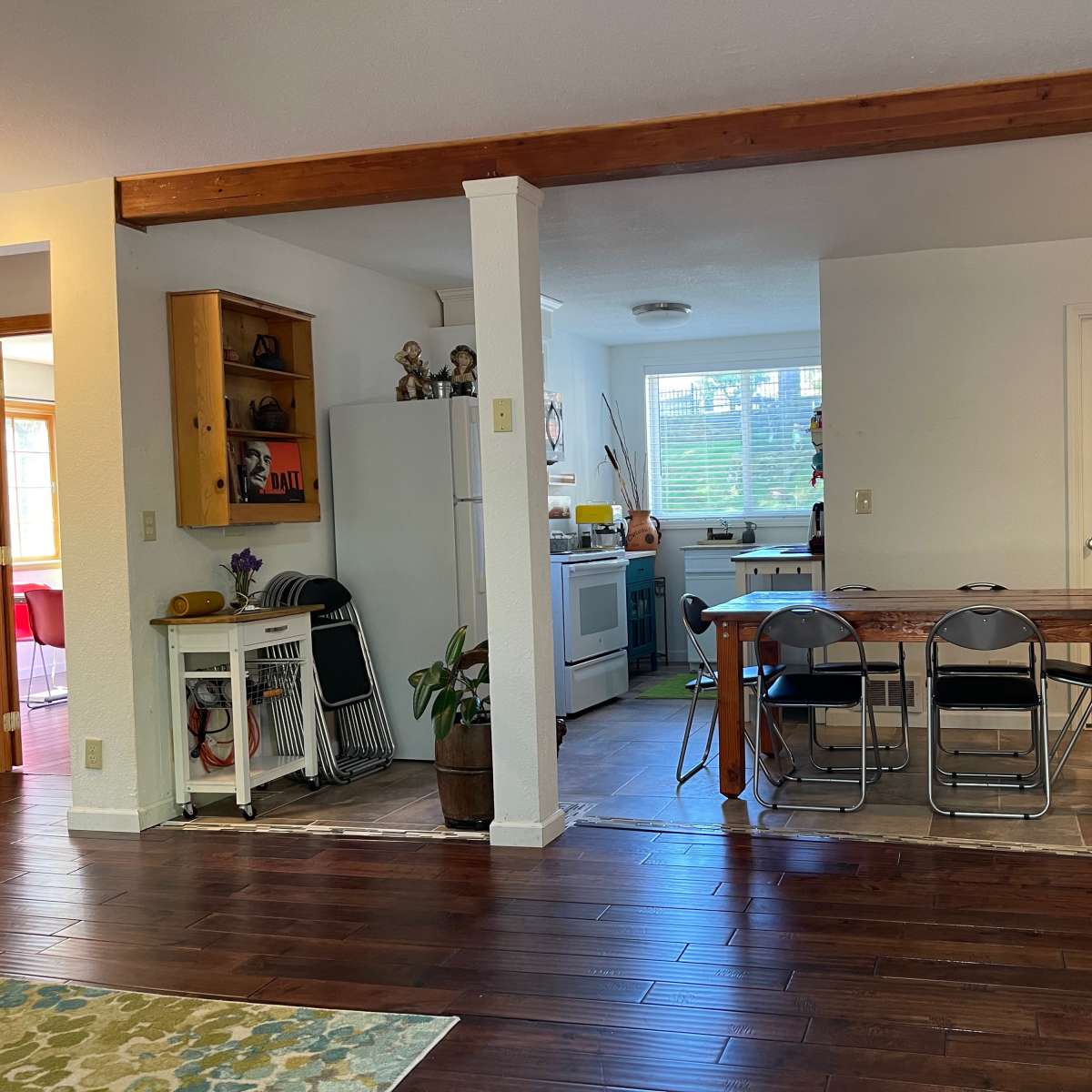 An open-concept kitchen and dining area with wooden beams, a dining table with metal chairs, and an adjoining kitchen featuring cabinets and a window.