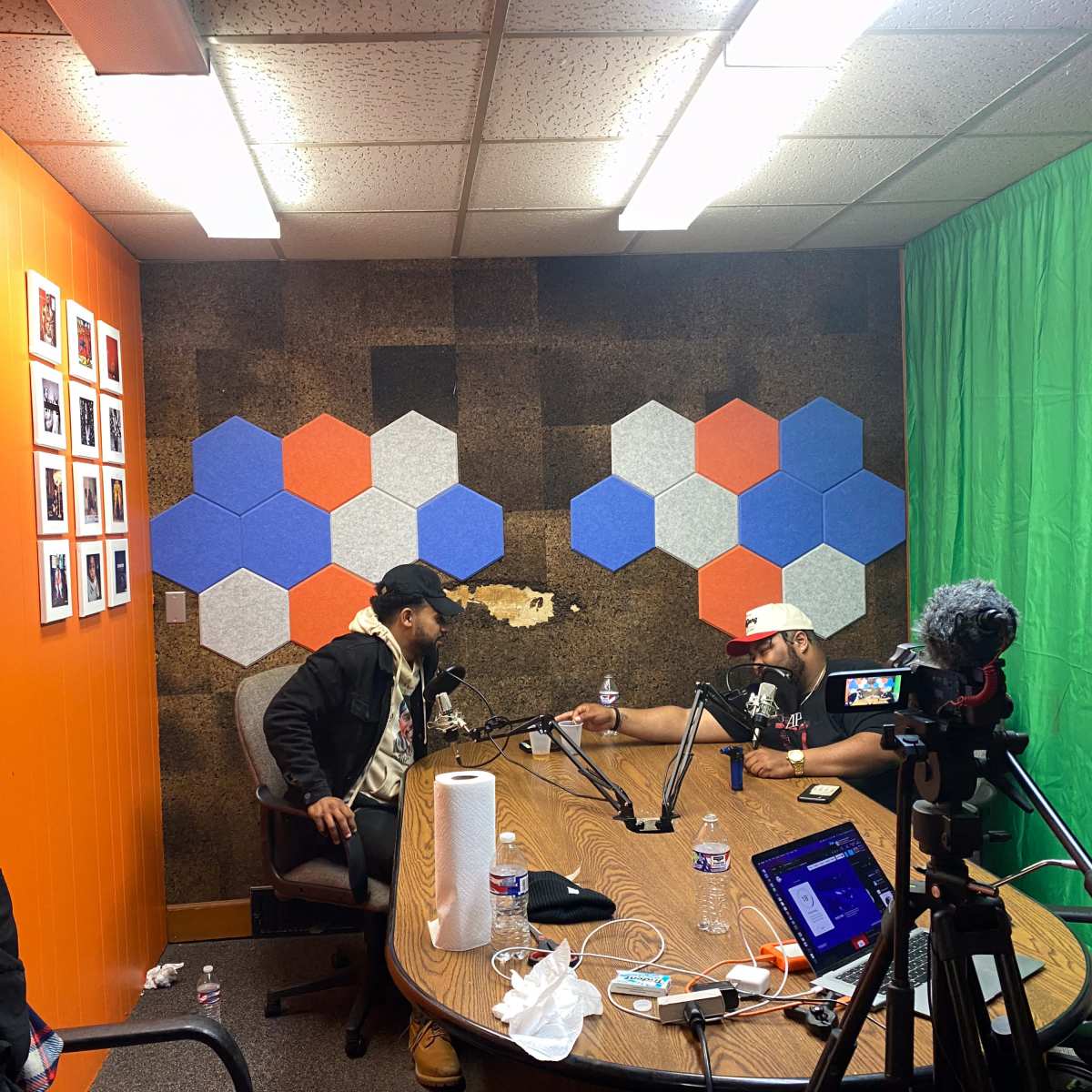 Two individuals are seated at a round table in a small studio with colorful hexagonal wall decorations and a green backdrop.