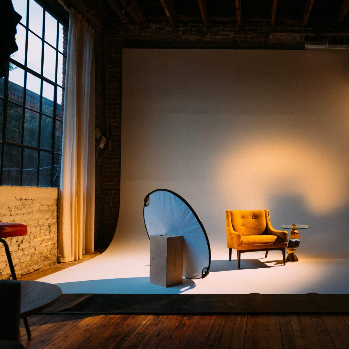 A yellow armchair positioned on a white backdrop in a studio, with a softbox light illuminating the scene and wooden flooring visible.