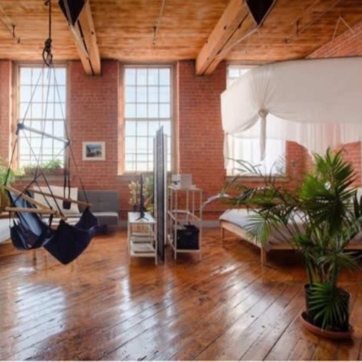 A spacious room with hardwood floors, large windows, a swing chair, and plants, featuring modern furniture against a brick wall.