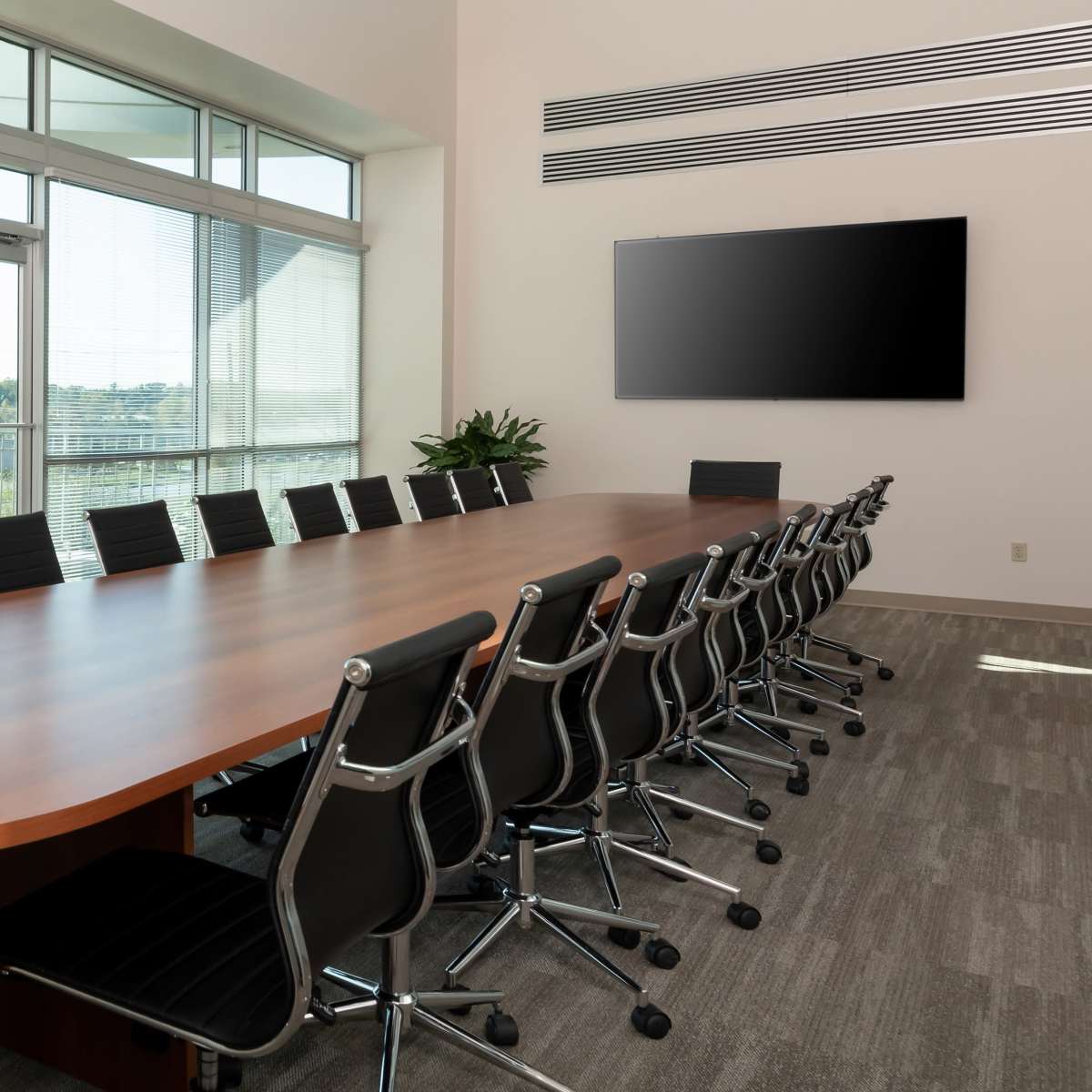 A large conference room features a long wooden table surrounded by black swivel chairs, with floor-to-ceiling windows letting in natural light and a wall-mounted television.