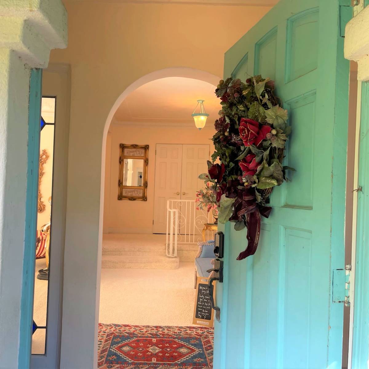 A turquoise door is open, revealing a bright hallway with a decorative mirror and a floral wreath adorning the door.