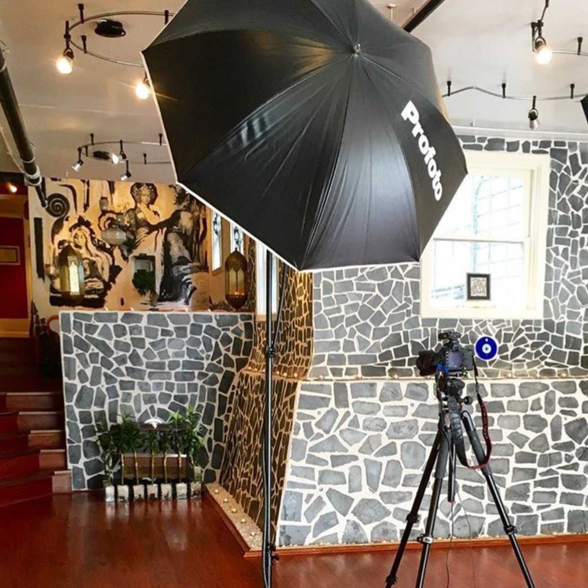 A photography setup features a black umbrella softbox and a camera on a tripod in a room with stone-patterned walls and wooden flooring.