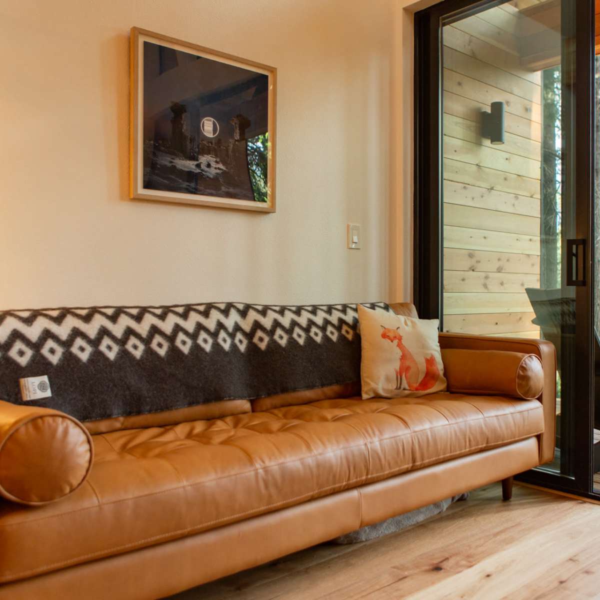 A leather sofa with a patterned throw pillow is positioned next to a large sliding glass door that opens to a wooden deck surrounded by trees.