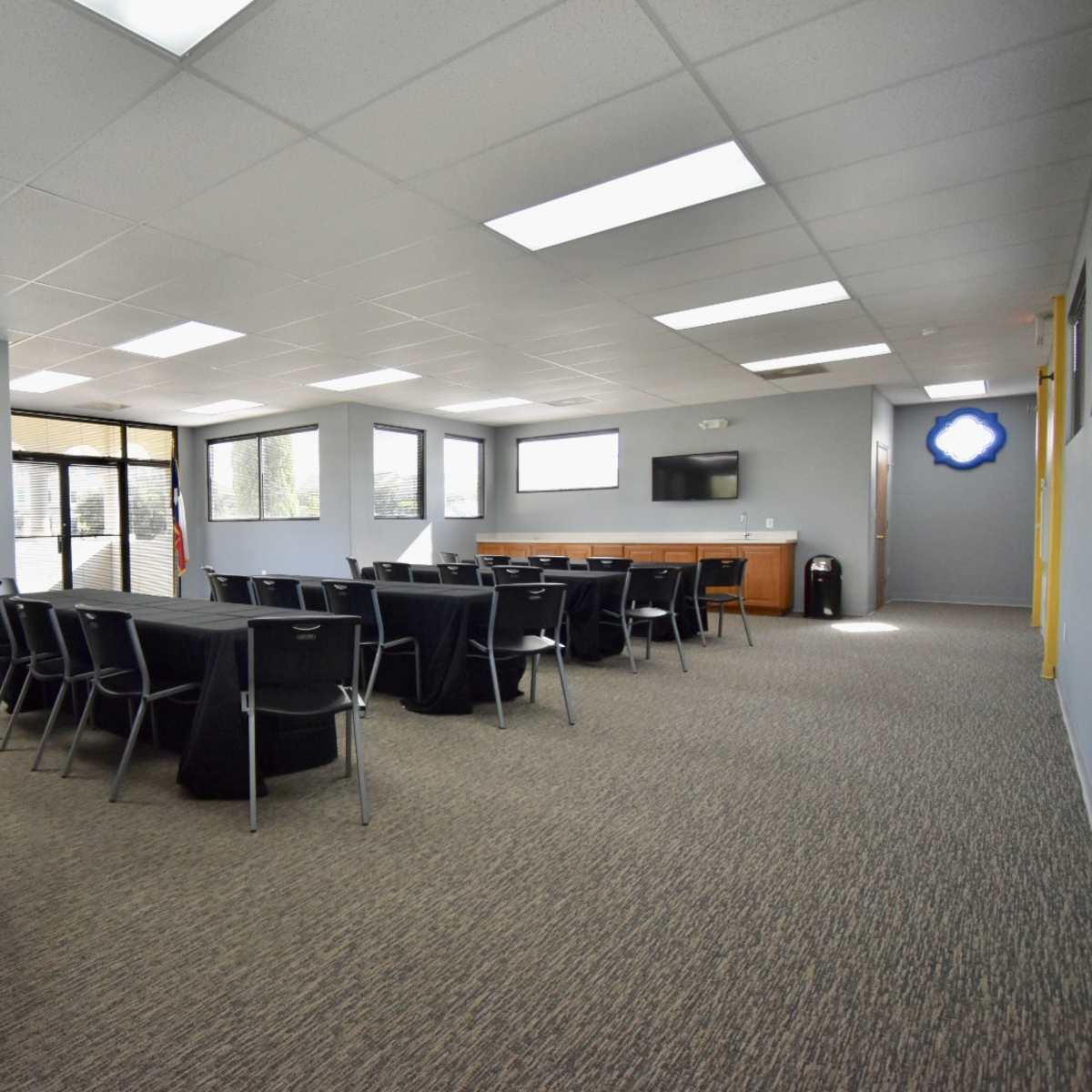 A conference room features multiple tables covered in black cloth, with chairs arranged neatly and large windows allowing natural light in.
