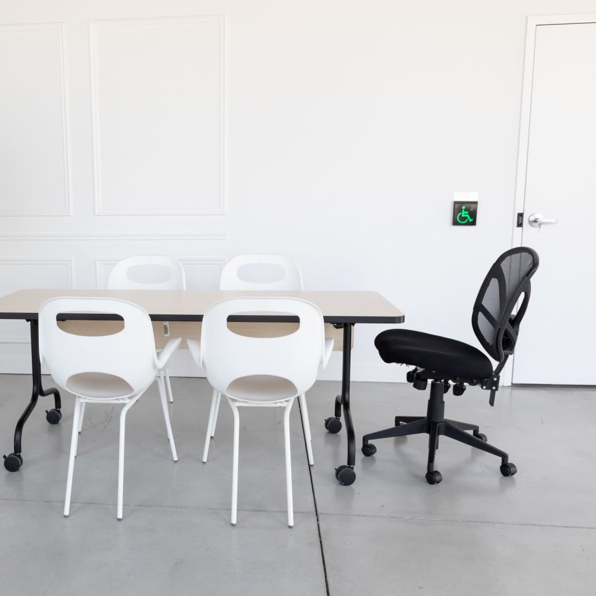 A meeting room features a rectangular table surrounded by four white chairs, with a black rolling office chair and a mounted television on a stand.