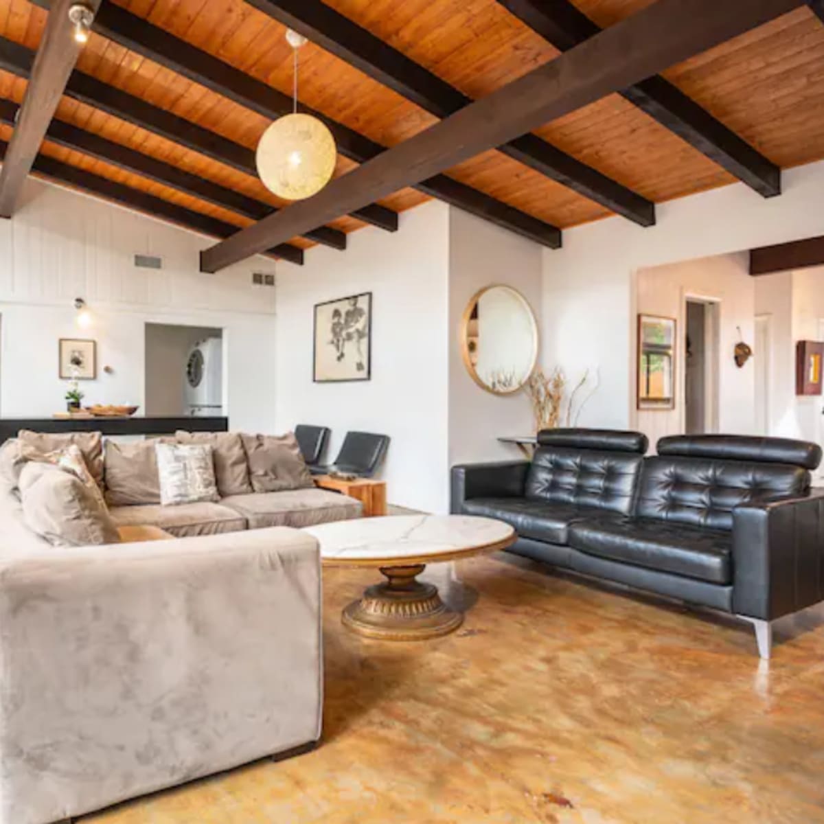 A modern living room with a high ceiling, exposed wooden beams, a beige sectional sofa, black leather chairs, and a round coffee table.