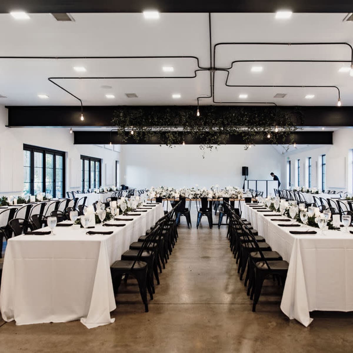 A banquet room with neatly arranged tables covered in white tablecloths, set for an event with centerpieces and chairs facing a stage area.