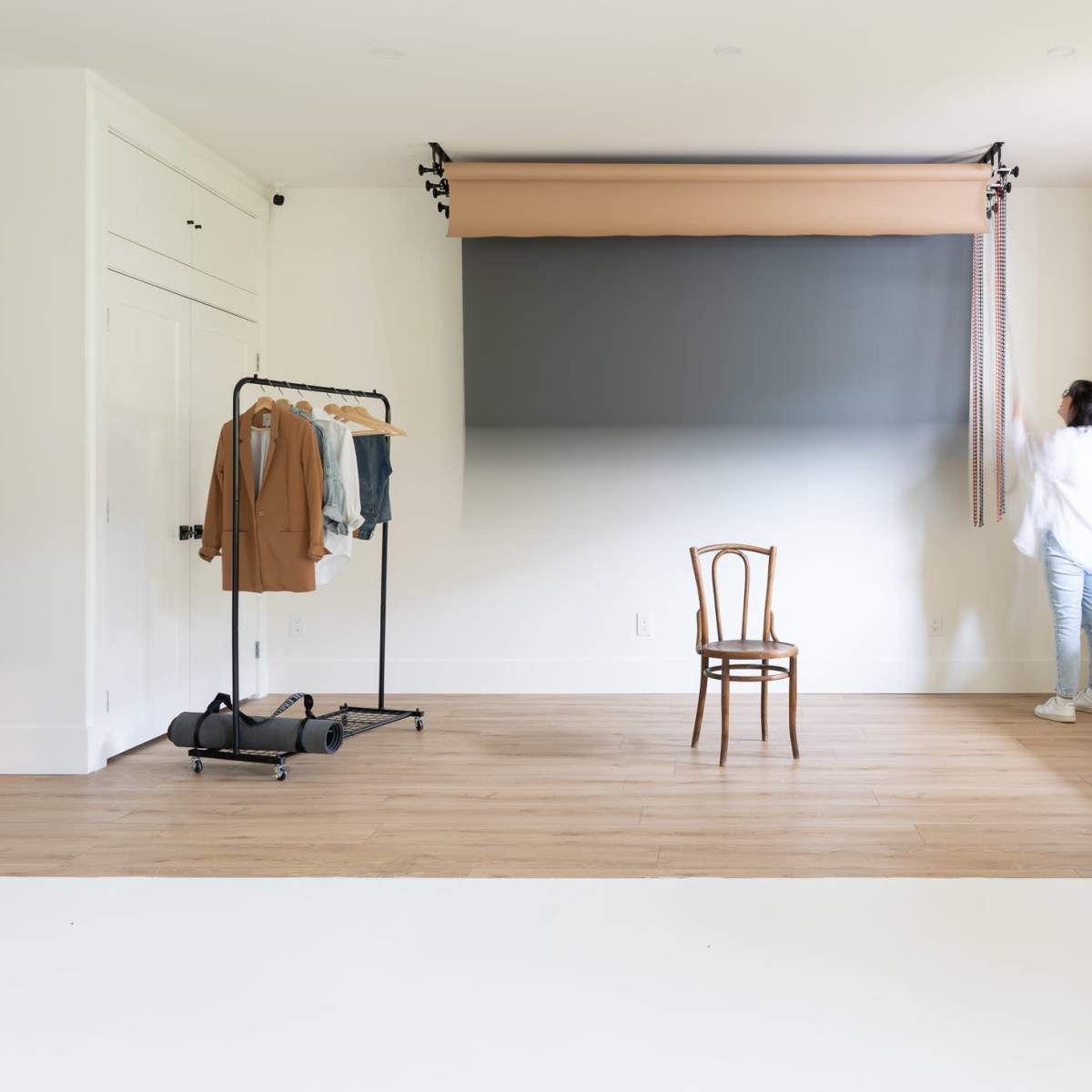A person is adjusting a fabric backdrop in a bright, minimalistic room with a clothing rack and a chair.