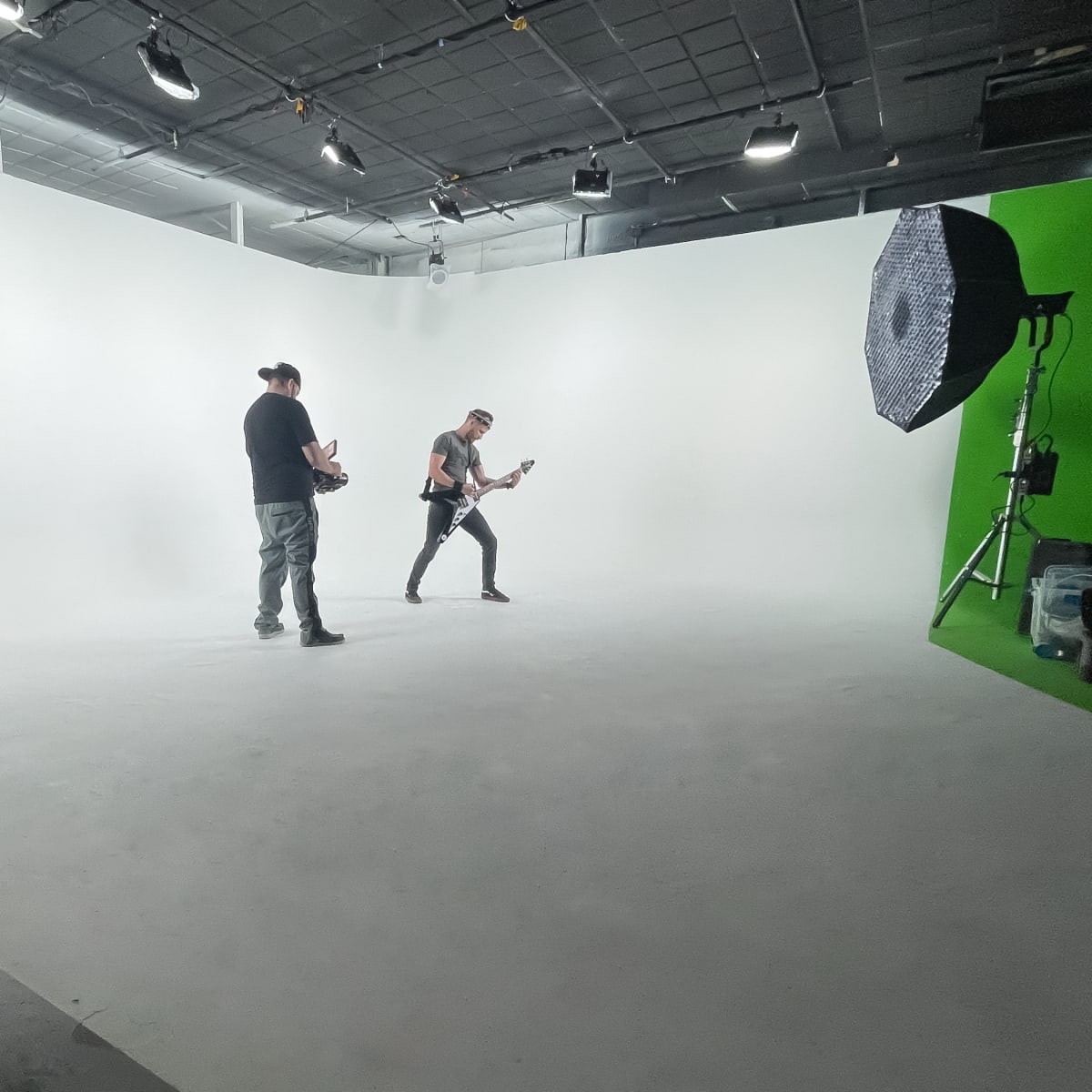 A musician is playing guitar on a white backdrop while another person stands nearby in a performance studio.