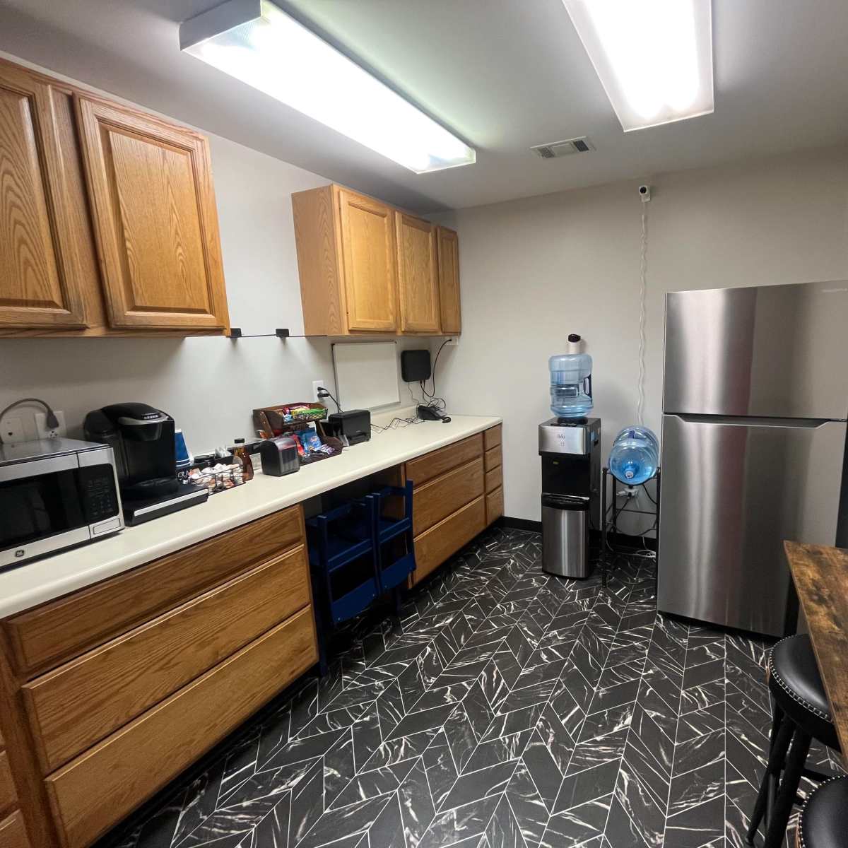 A kitchen with wooden cabinets, a microwave, a coffee maker, a stainless steel refrigerator, a water cooler, and a separate dining table with two black stools.