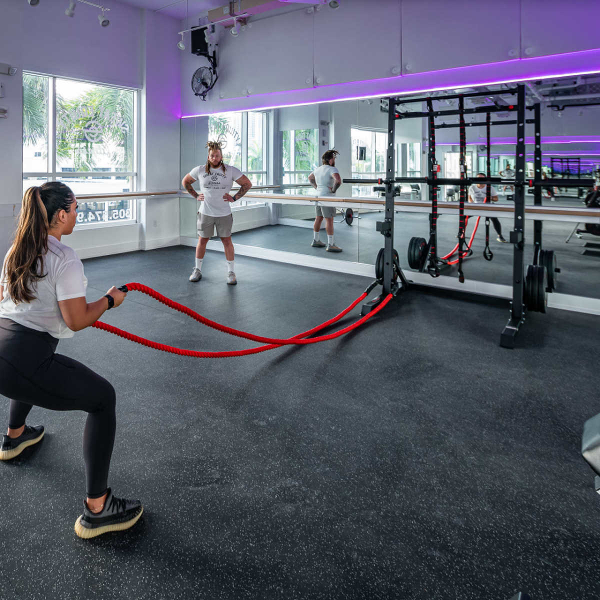 A woman performs a battle rope exercise while a trainer observes in a modern gym space.