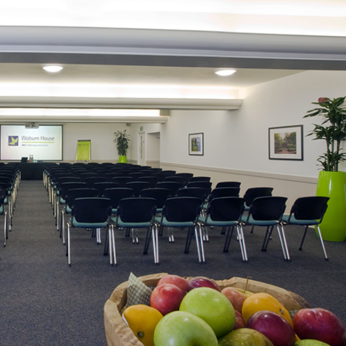 A large, empty conference room with rows of black chairs facing a presentation screen, complemented by a table of pastries and fruit in the foreground.