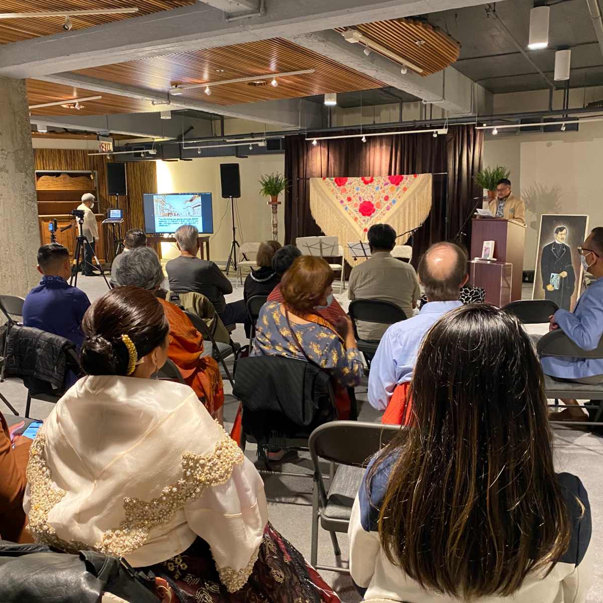 A diverse audience is seated in a spacious event room, where a speaker addresses them from a podium near a decorative backdrop.