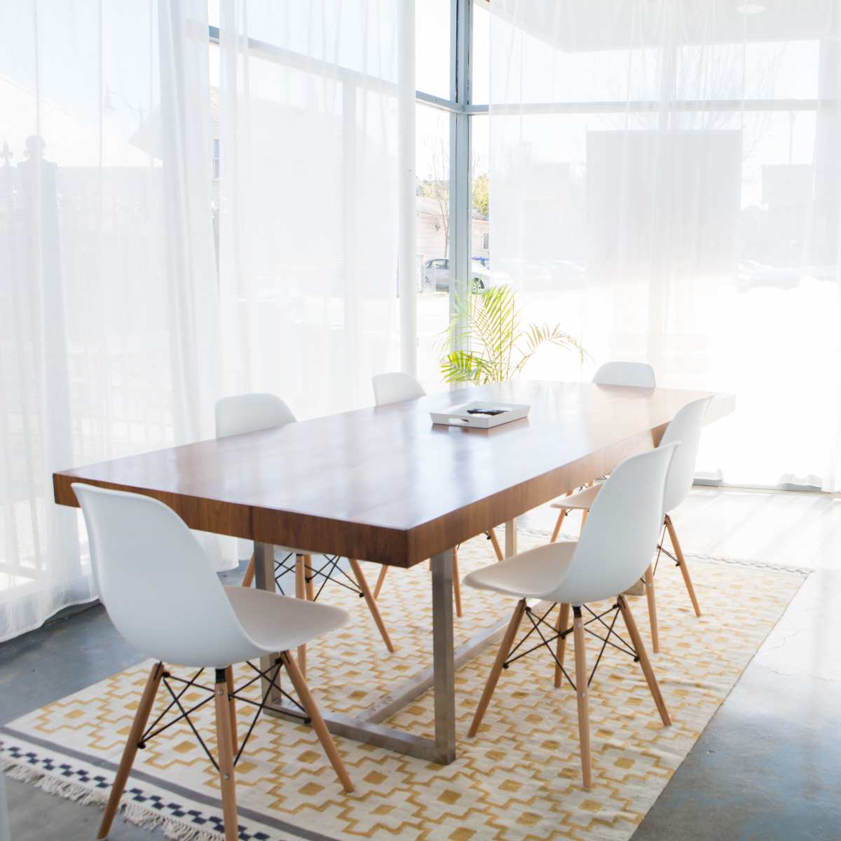 A modern conference room features a wooden table surrounded by white chairs, with large windows partially covered by sheer curtains.