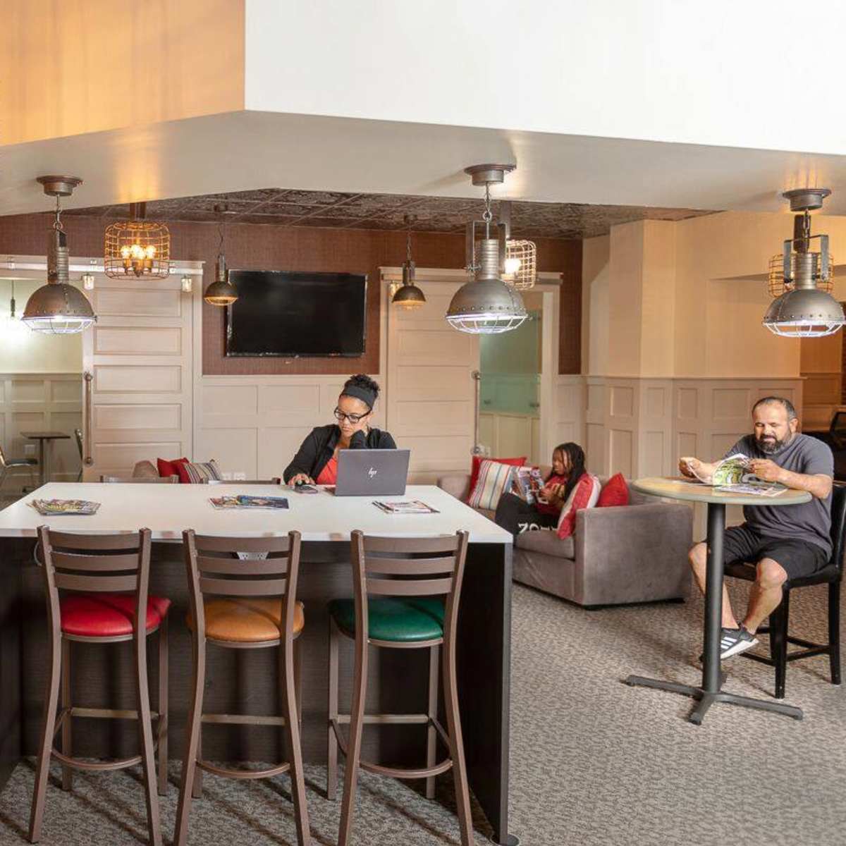 A group of four people work on laptops at a modern common area featuring bar stools, pendant lighting, and a television on the wall.