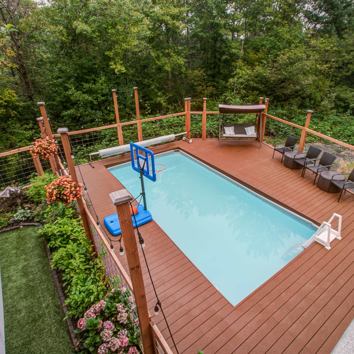 A rectangular swimming pool surrounded by a wooden deck, seating chairs, and lush greenery in the background.