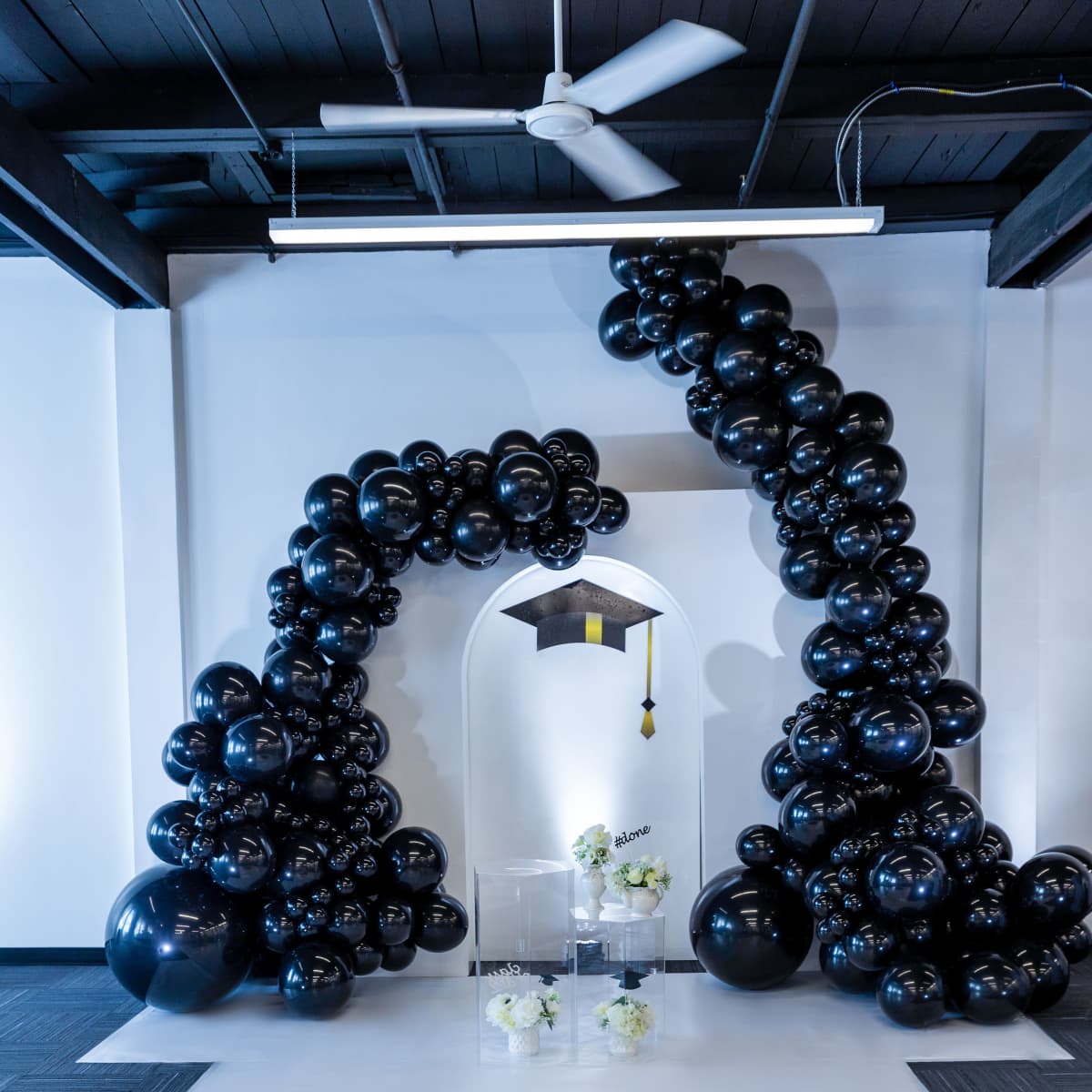 A decorative arch made of black balloons with a graduation cap at the top, set against a white wall, with a sign reading "Congratulations" on the floor.
