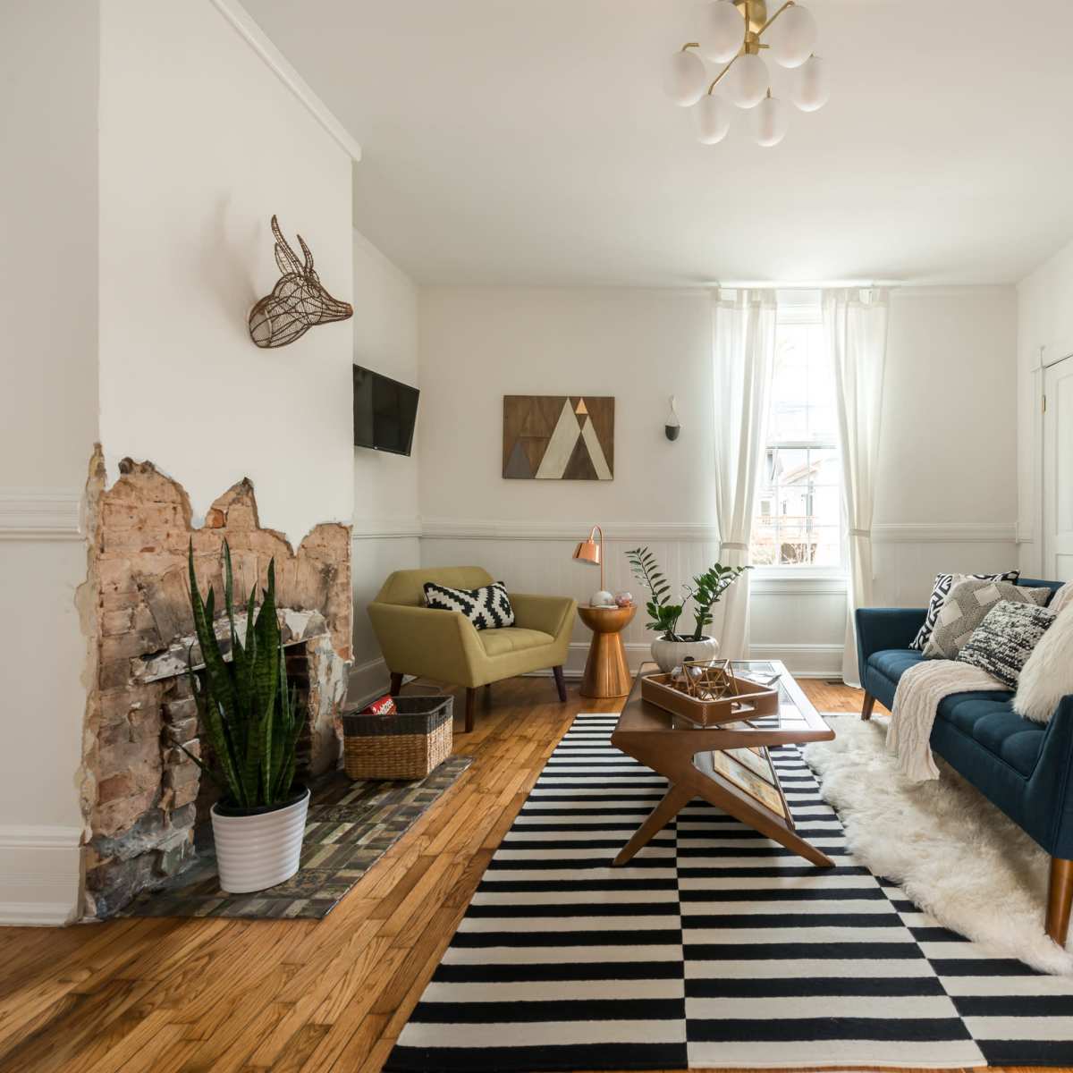 The living room features a blue sofa, a green armchair, a wooden coffee table, and a patterned black and white rug, with a partially exposed brick wall and large windows allowing natural light.