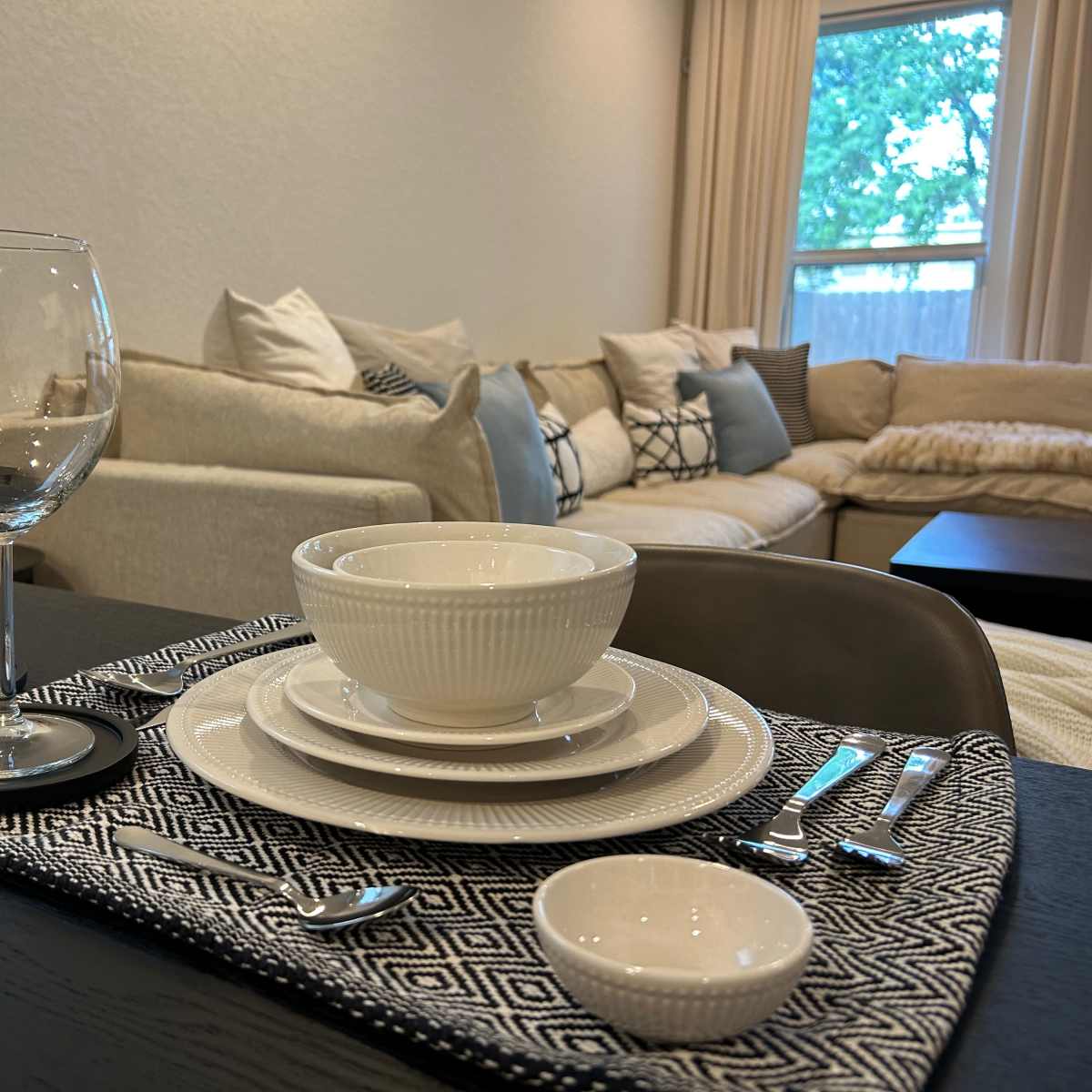 A neatly arranged dining table setting features white dinnerware, glassware, and cutlery, with a cozy living area in the background.
