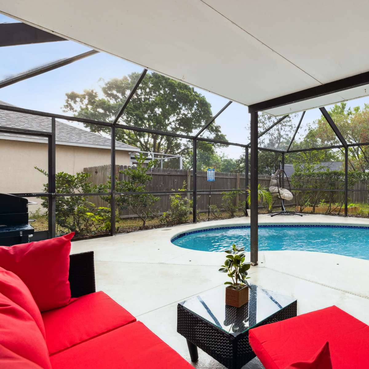 A covered patio features red seating and a small table beside a rectangular pool surrounded by a screened enclosure and landscaped yard.