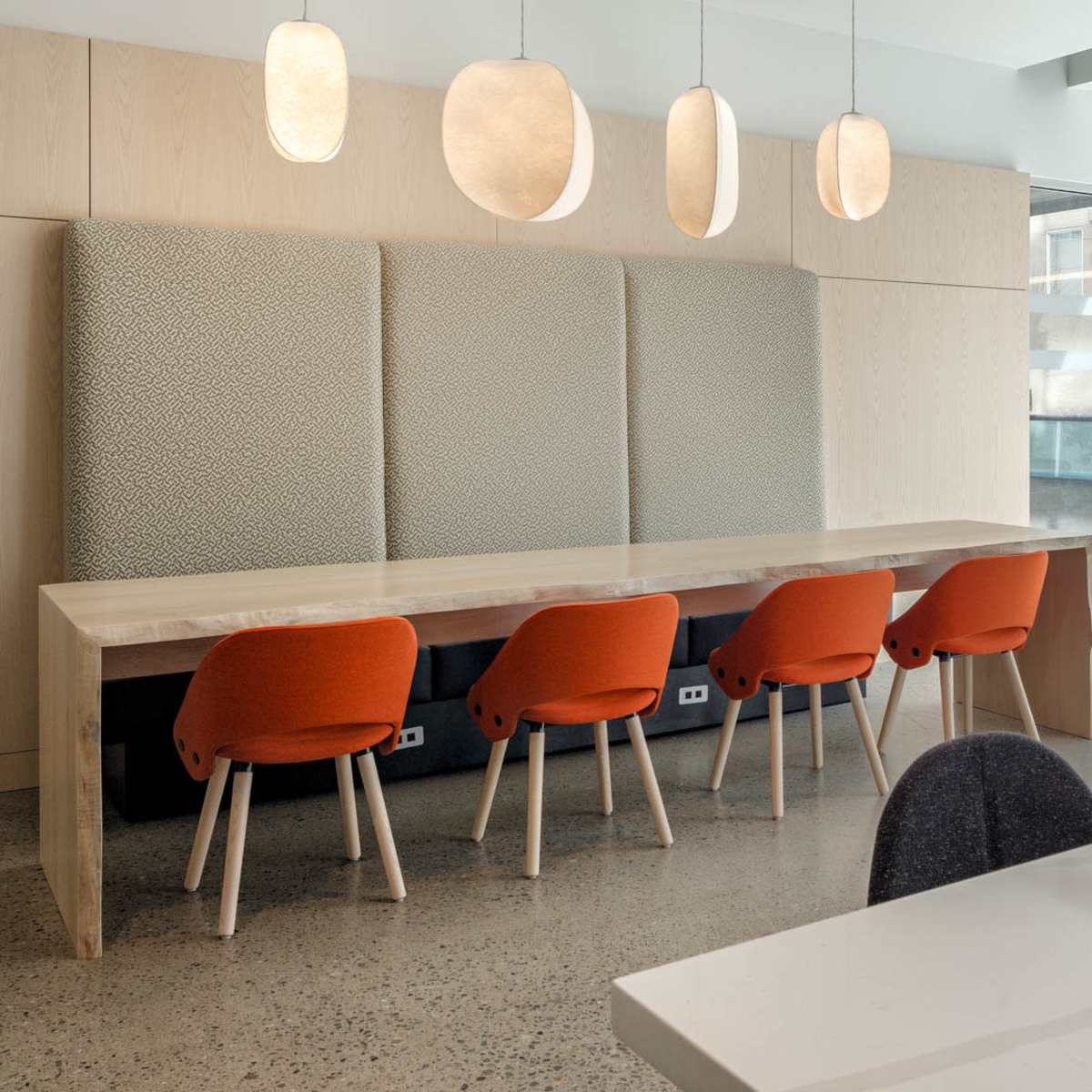 A modern dining area features a long wooden table with five orange chairs, complemented by pendant lights and a textured wall backdrop.