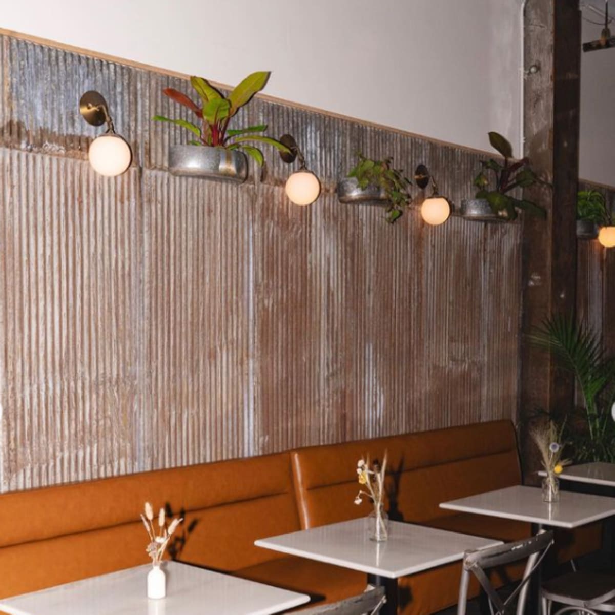 A modern cafe interior with metal and wooden accents featuring a row of small tables and orange seating along a textured wall.
